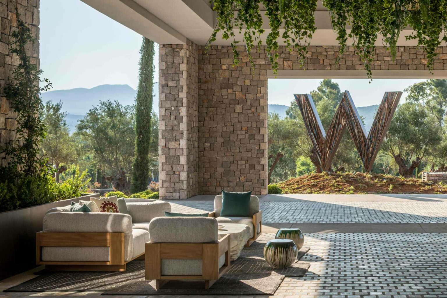Spacious hotel lounge area with stone walls, beige sofas, and green pillows, overlooking a scenic landscape with trees and a large 