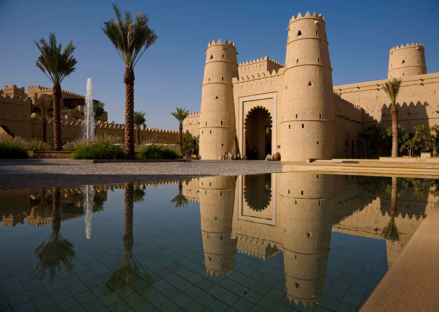 A historic sand-colored fort with tall, cylindrical towers reflects in a tranquil pool. Palm trees and a spouting fountain enhance the serene scene.