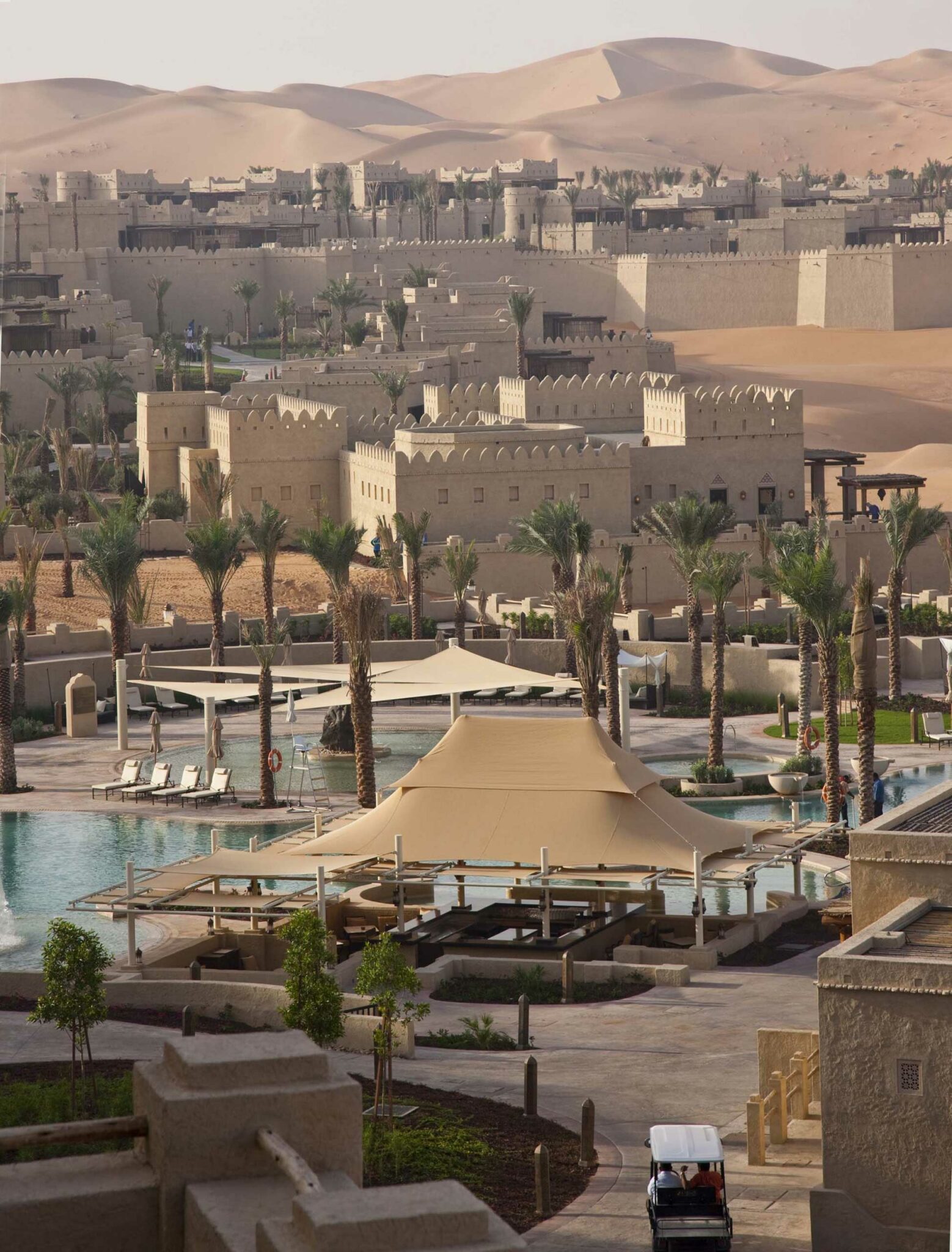 The luxurious Qasr Al Sarab Desert Resort by Anantara with tan buildings and palm trees against rolling sand dunes. A central pool is shaded by large beige canopies, creating a tranquil oasis.