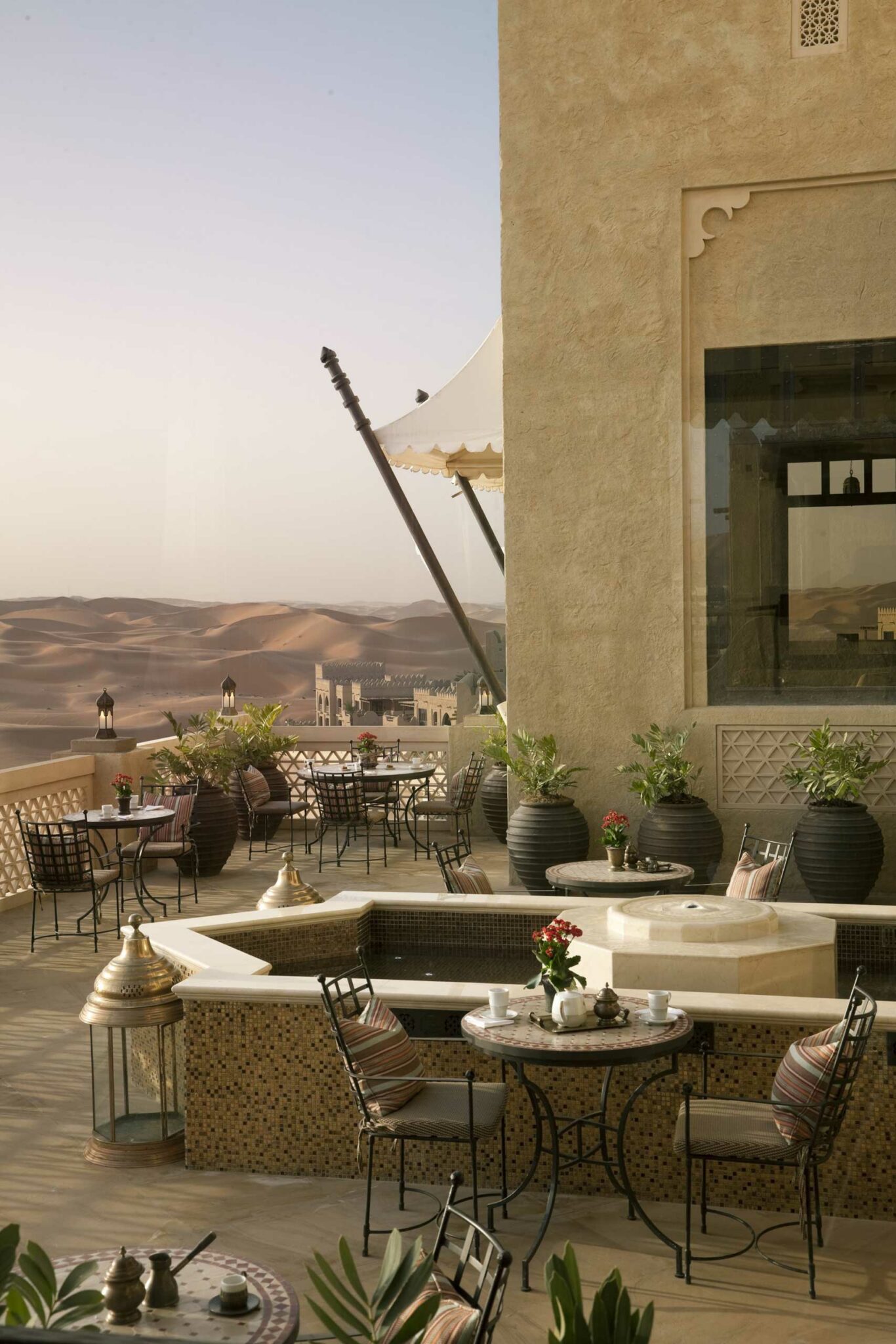 Luxurious desert resort terrace with ornate tables, potted plants, and lanterns overlooks vast dunes at sunset, evoking a serene and elegant ambiance.