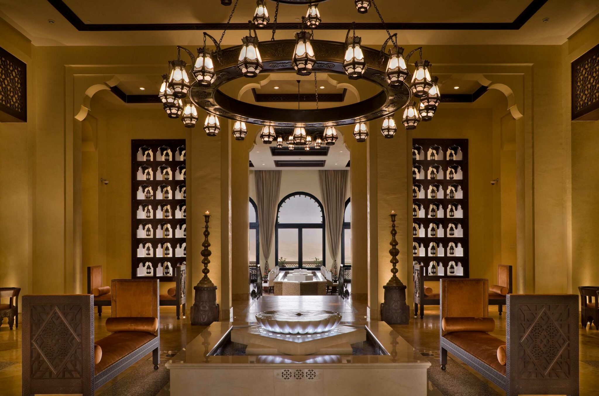 Luxurious resort interior with warm lighting. Elegant chandelier hangs above a central water feature. Arched windows and shelves create a sophisticated ambiance.