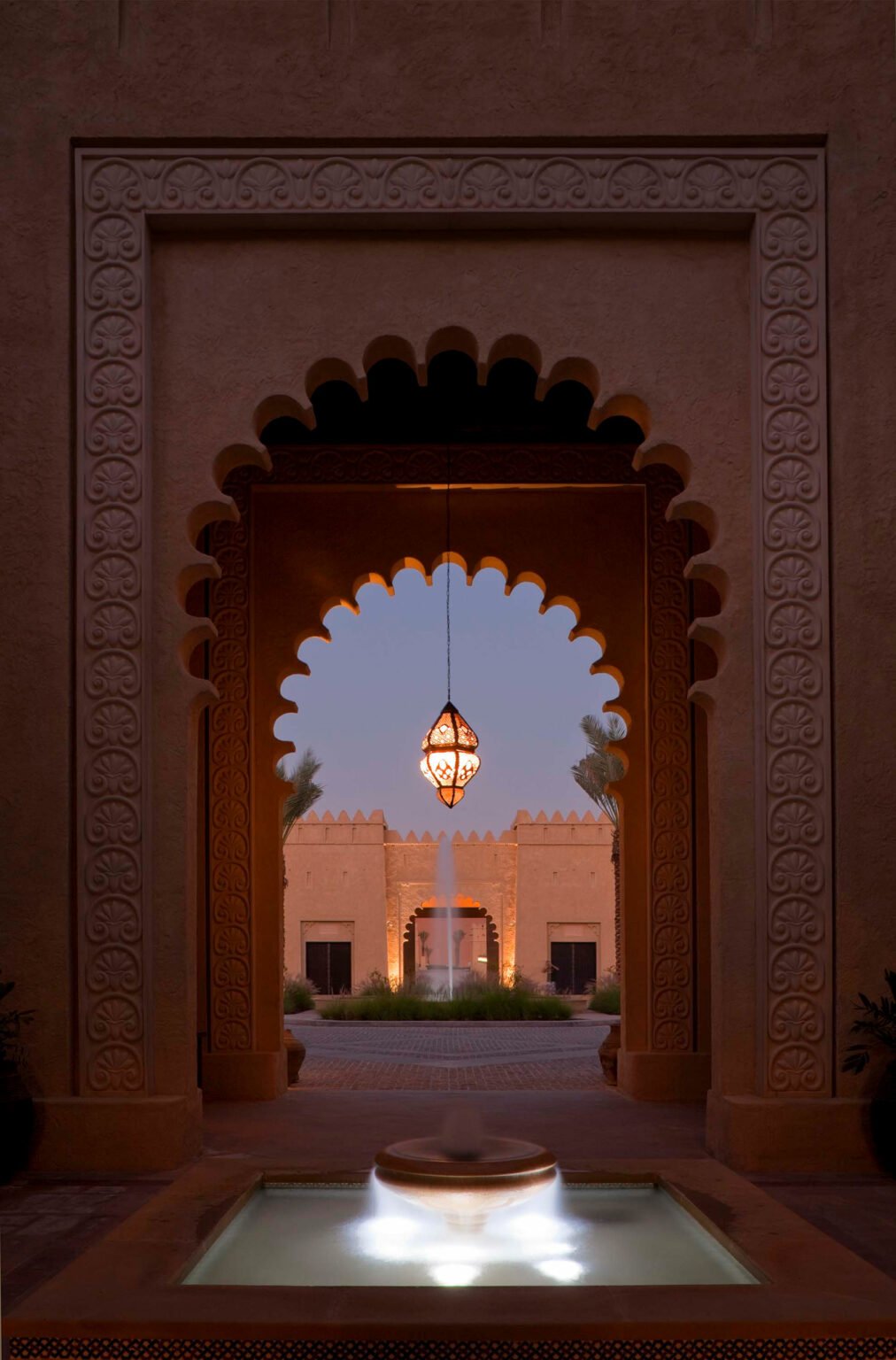 Ornate archway frames a glowing lantern and serene fountain, leading to a softly lit courtyard with palm accents, evoking a tranquil, warm ambiance.