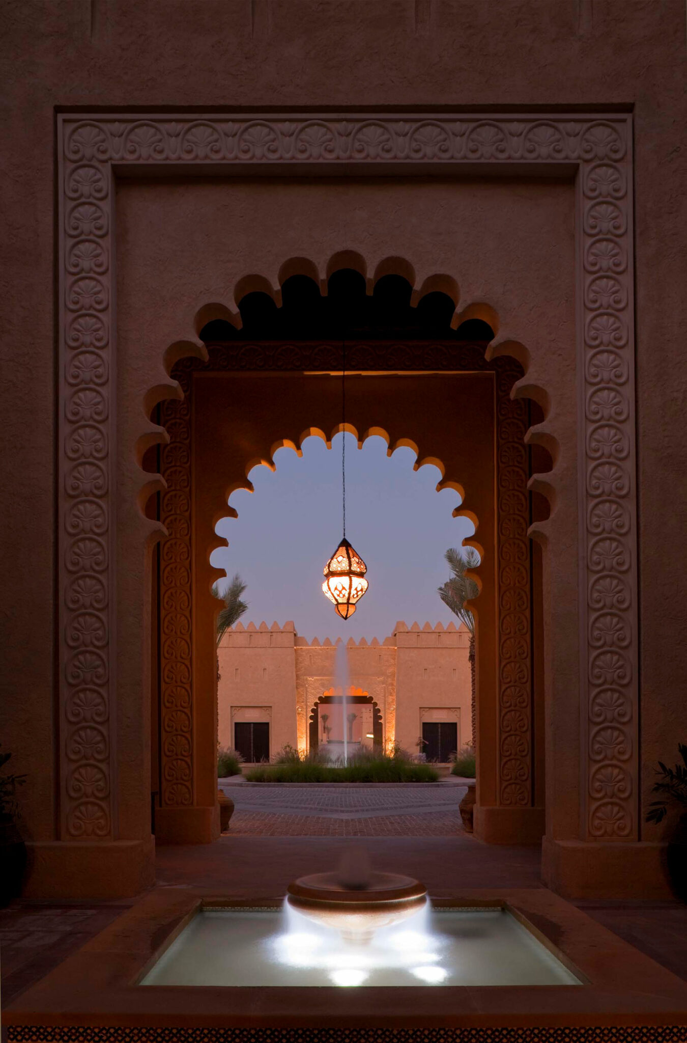 Ornate archway frames a glowing lantern and serene fountain, leading to a softly lit courtyard with palm accents, evoking a tranquil, warm ambiance.