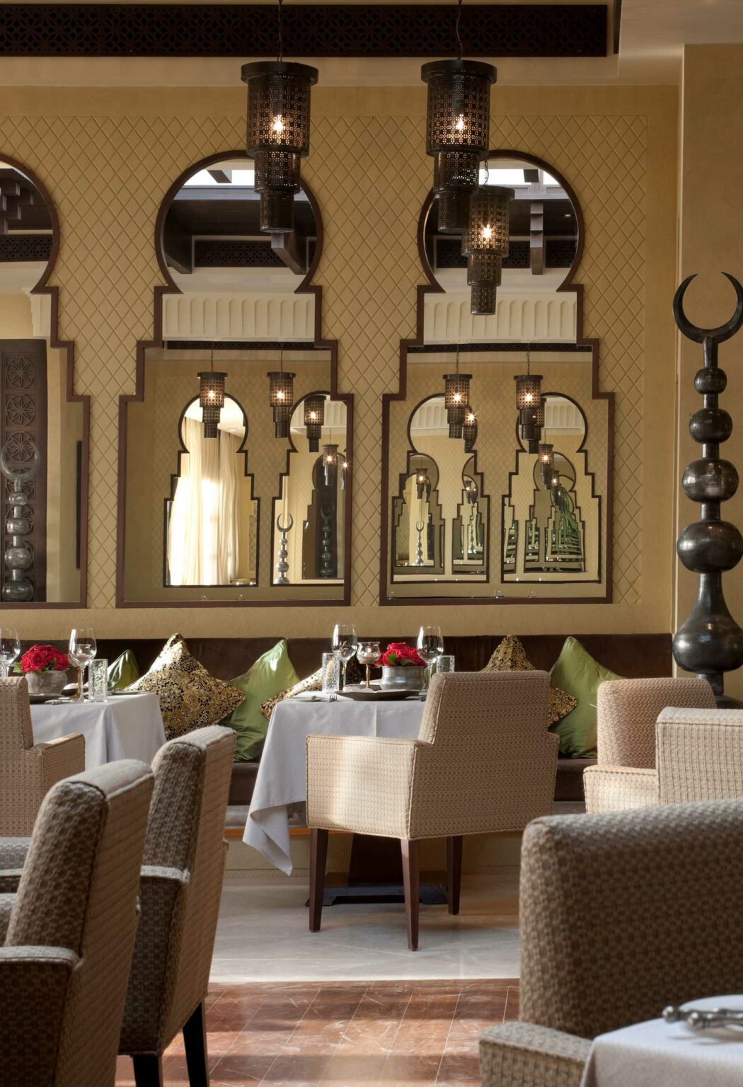 Elegant resort restaurant interior with plush chairs and white tablecloths. Ornate mirrors and hanging lanterns reflect warm ambient lighting, creating a cozy atmosphere.