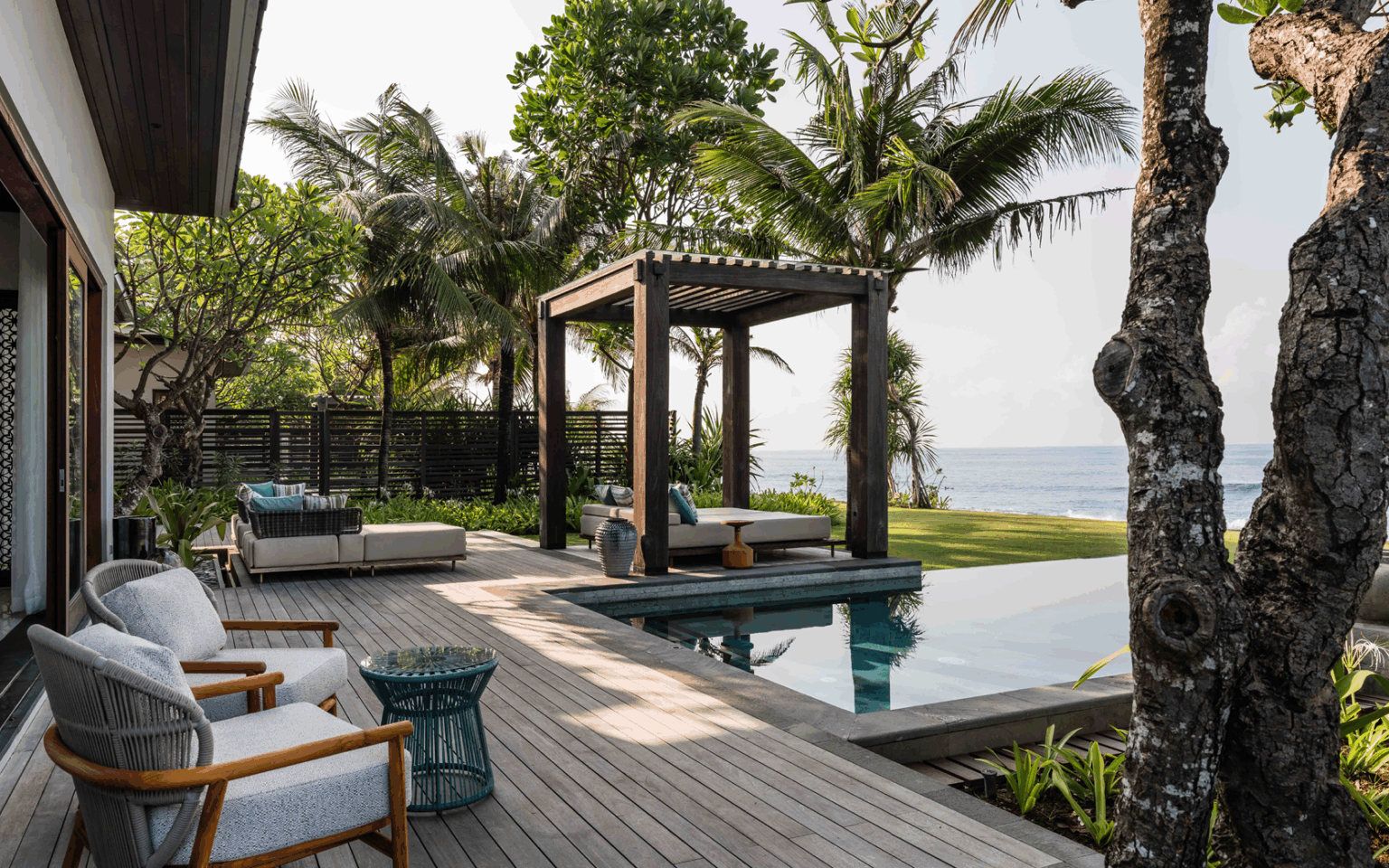 Tropical poolside oasis with wooden deck, lounge chairs, and a pergola. Palm trees and ocean view create a serene, relaxing atmosphere.