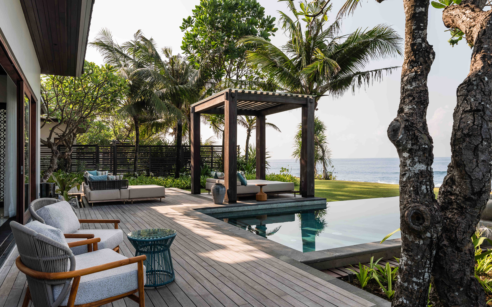 Tropical poolside oasis with wooden deck, lounge chairs, and a pergola. Palm trees and ocean view create a serene, relaxing atmosphere.