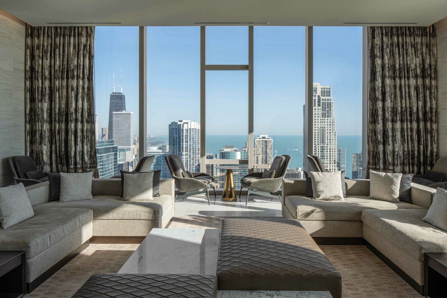 Luxurious living room with beige sofas and cushions, large windows offer a city skyline and lake view. Sunlight brightens the elegant space.