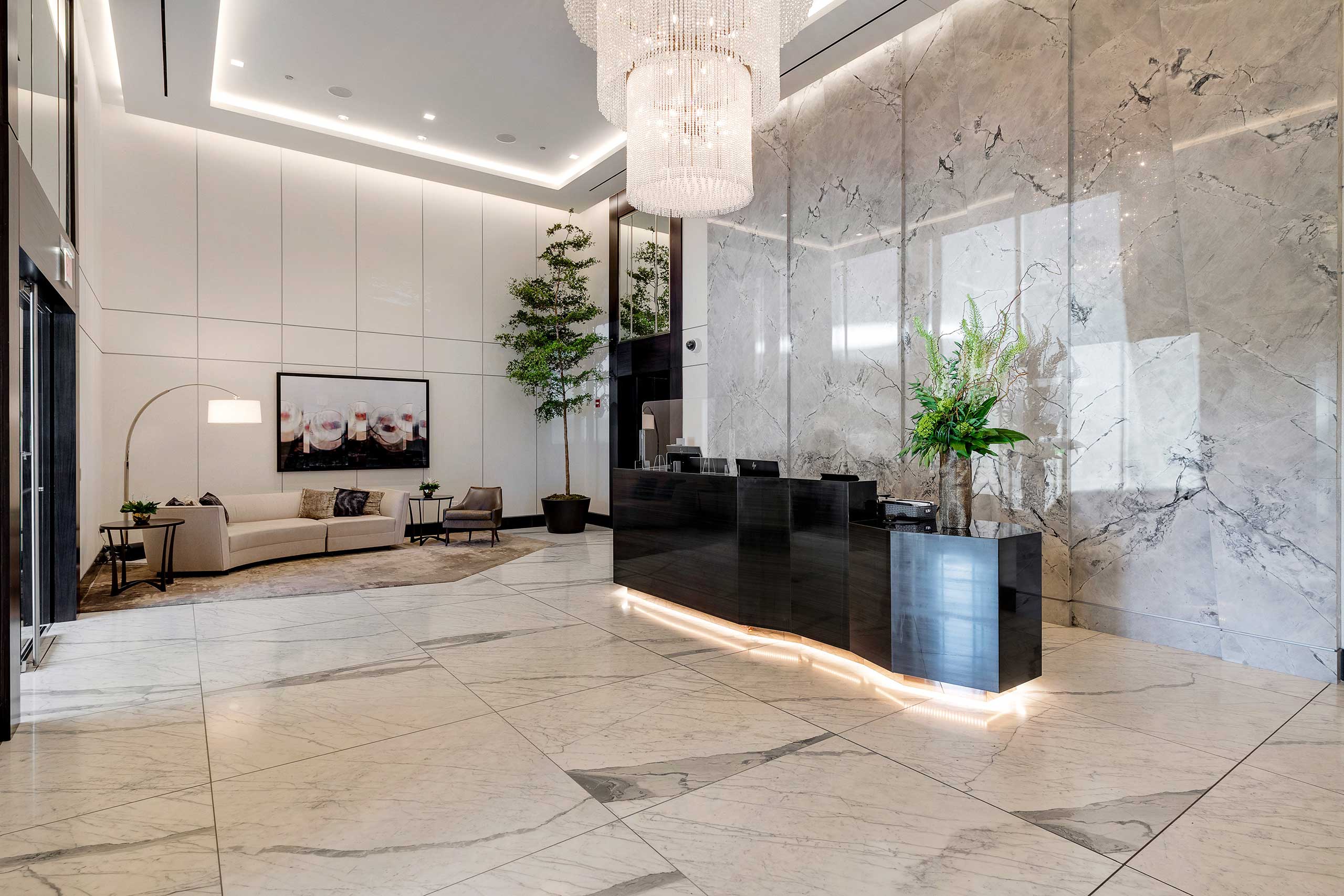 Luxurious residential lobby with a marble floor and walls, elegant black reception desk, large chandelier, indoor plants, sofa, and wall art creating a sophisticated ambiance.