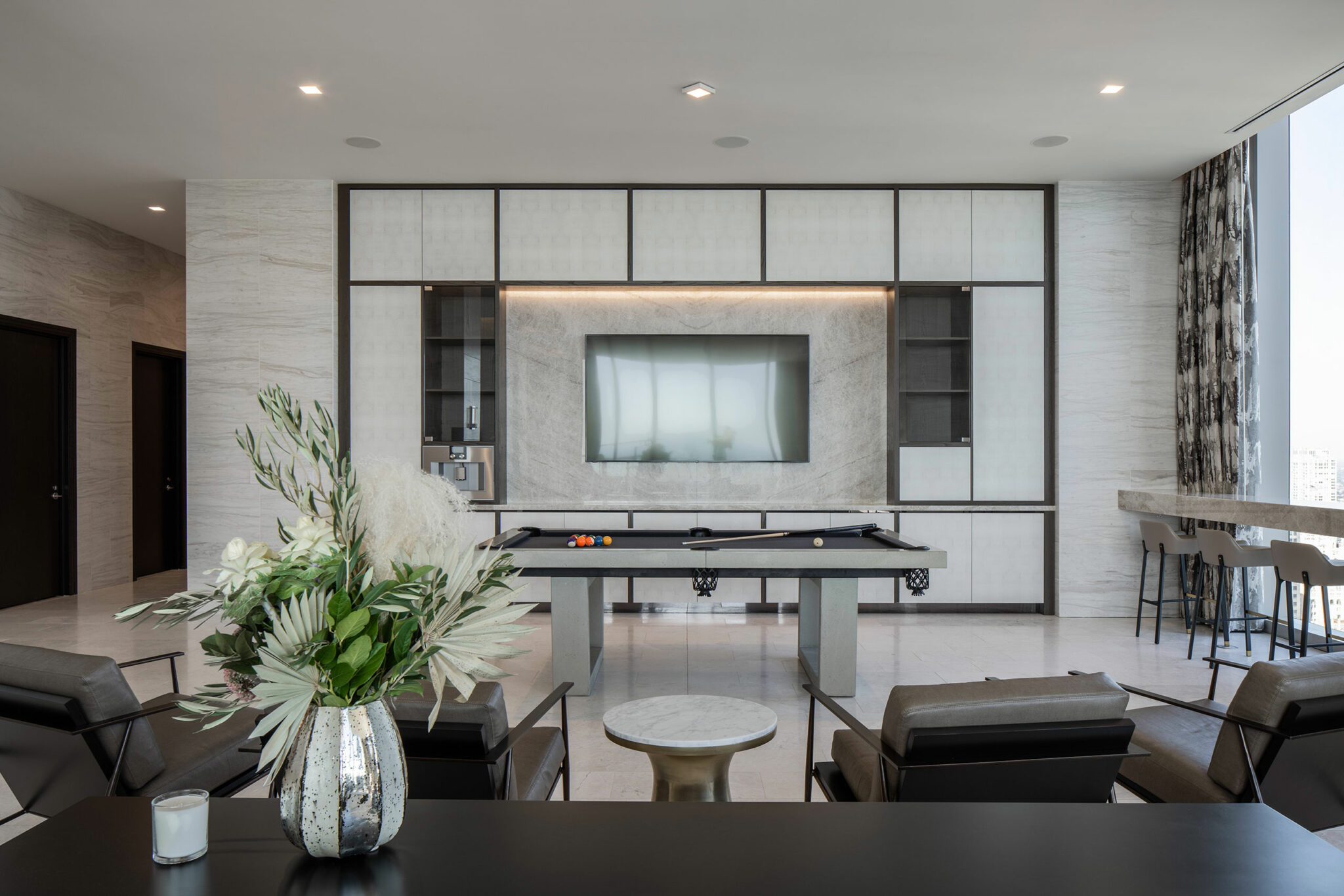 Modern home lounge area with a sleek pool table, wall-mounted TV, and elegant seating. A vase of white flowers adds sophistication. Bright, spacious atmosphere.