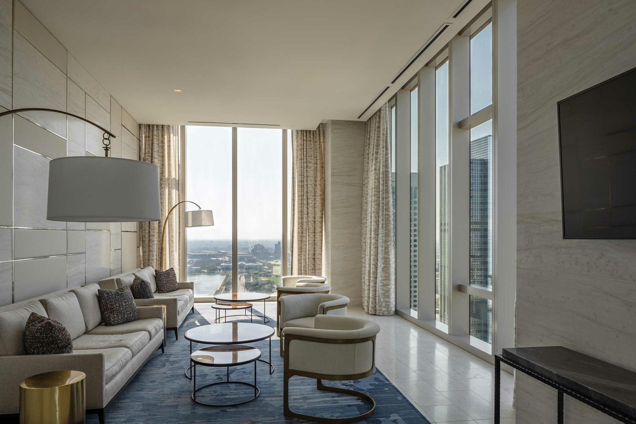 Luxurious living room with a modern beige sofa, accent chairs, and large windows offering a cityscape view. The atmosphere is serene and elegant.