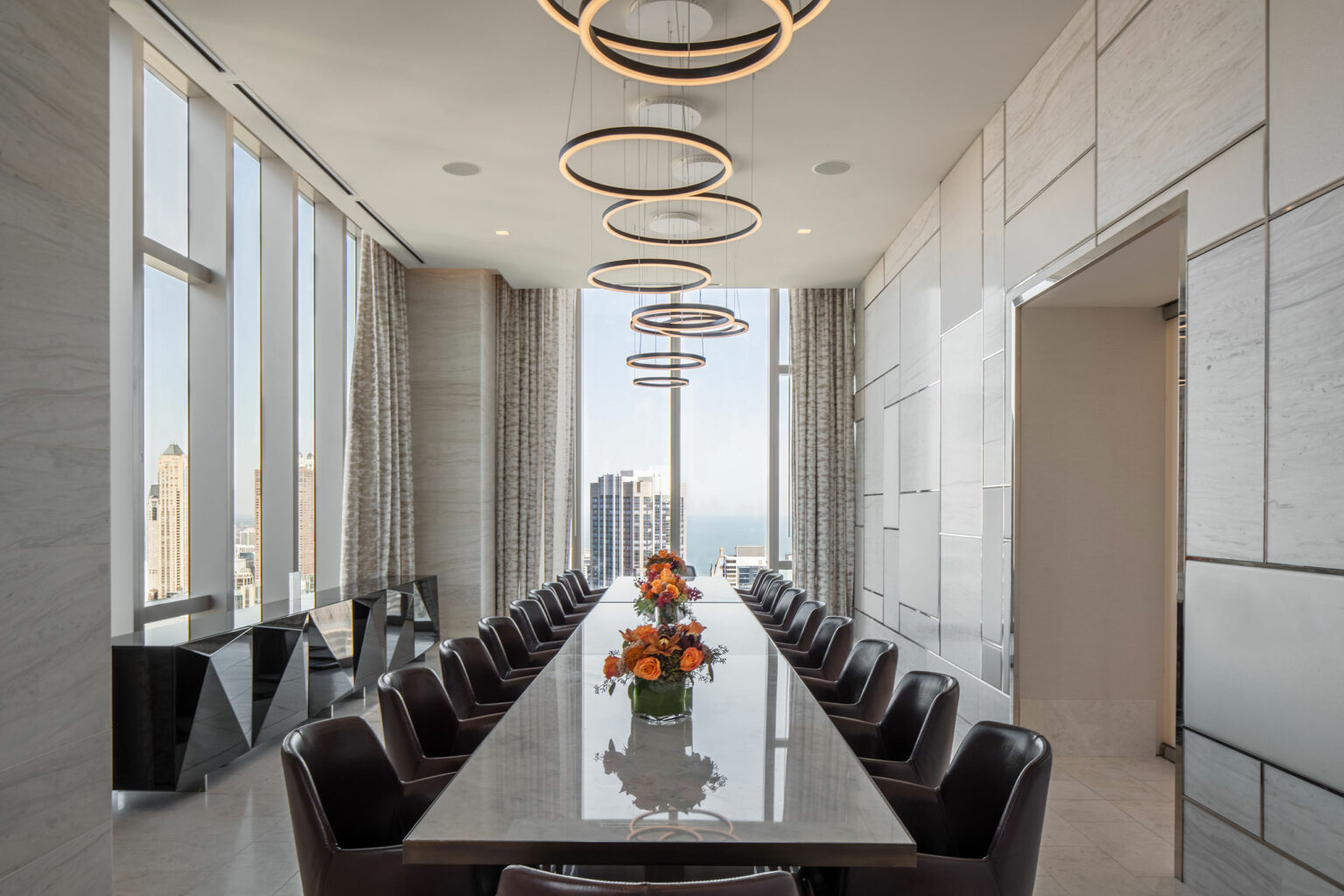 A sleek home conference room features a long, reflective table with modern chairs, framed by tall windows. Hanging circular lights and a floral centerpiece add elegance.