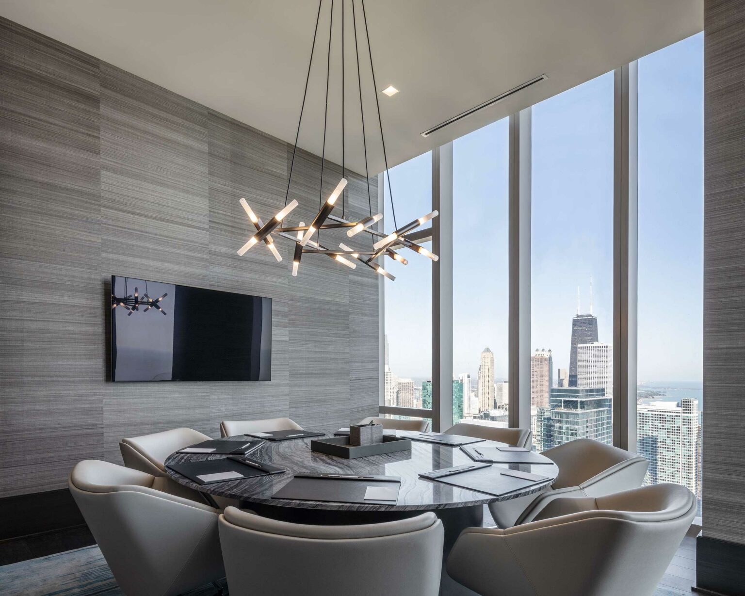 Modern home conference room with a large round marble table, sleek chairs, and geometric chandelier. Floor-to-ceiling windows reveal a city skyline.