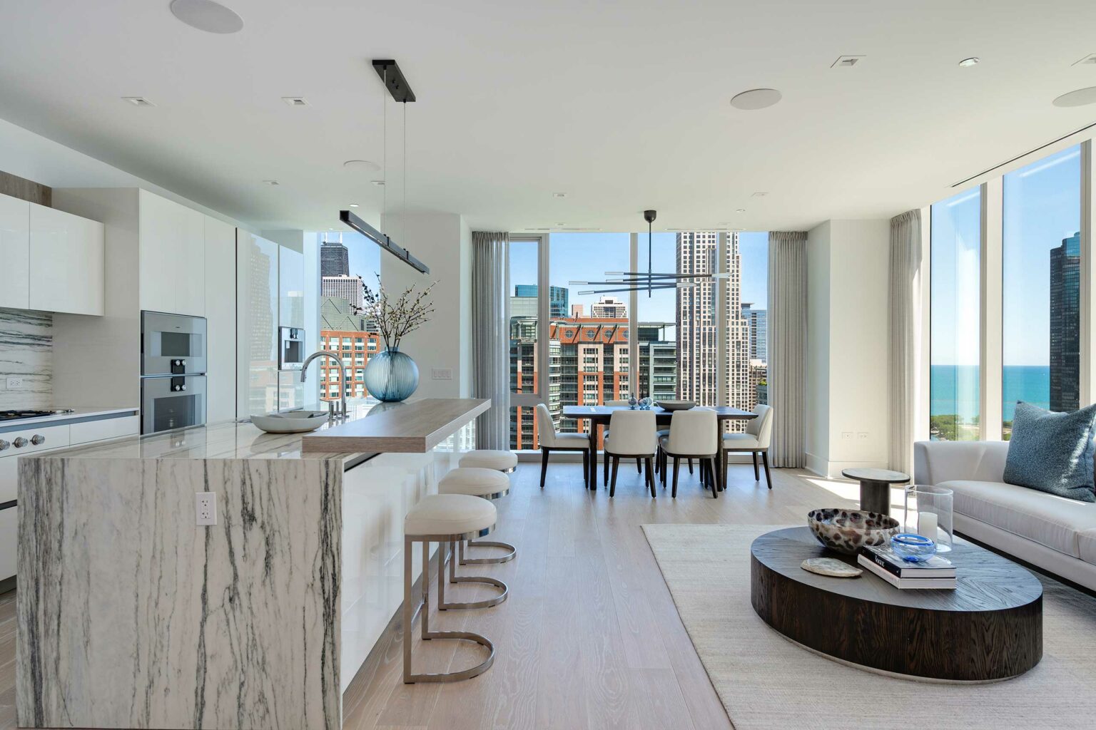 Modern open-plan living area with a marble kitchen island, elegant dining table, and cozy seating. Large windows reveal a cityscape and lake view.