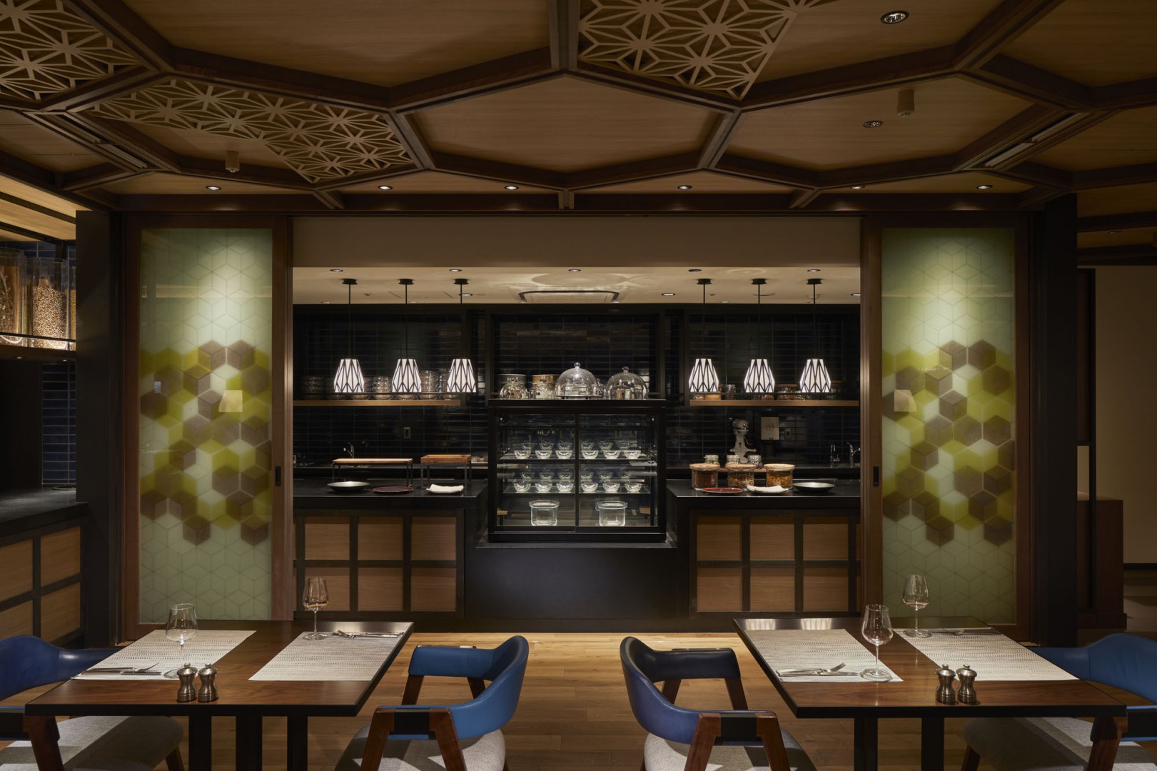 Elegant restaurant interior with a warm, cozy atmosphere. Features honeycomb-patterned glass panels, geometric ceiling, and sleek lighting over tables.