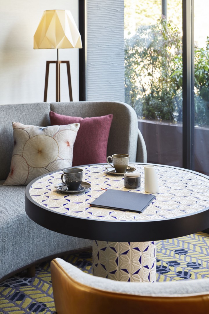 Cozy cafe area of a restaurant with a round, patterned table holding three cups and a notebook. Gray sofa with colorful pillows, geometric lamp, and window view.