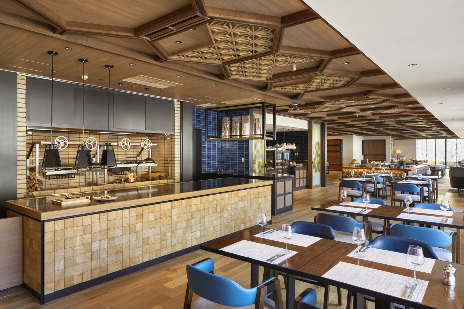 Modern restaurant interior with wooden ceiling and tiled walls, featuring a sleek, open kitchen. Rows of tables set with blue chairs create a welcoming ambiance.