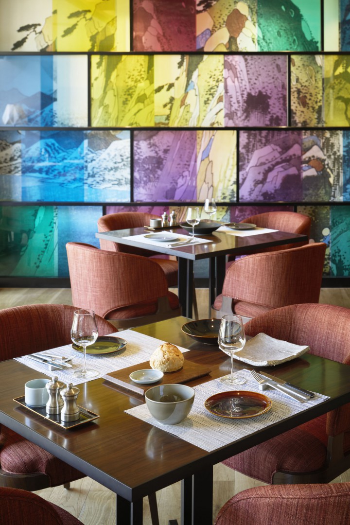 A modern restaurant with colorful abstract wall art. Two tables are set with wine glasses, plates, and utensils. Warm tones create an inviting atmosphere.