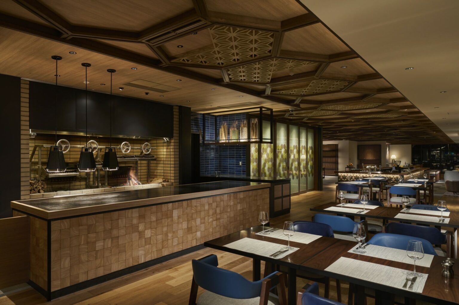 Modern restaurant interior with wooden decor and geometric ceiling patterns. Blue chairs surround tables set with placemats, glasses, and cutlery. Warm, ambient lighting.