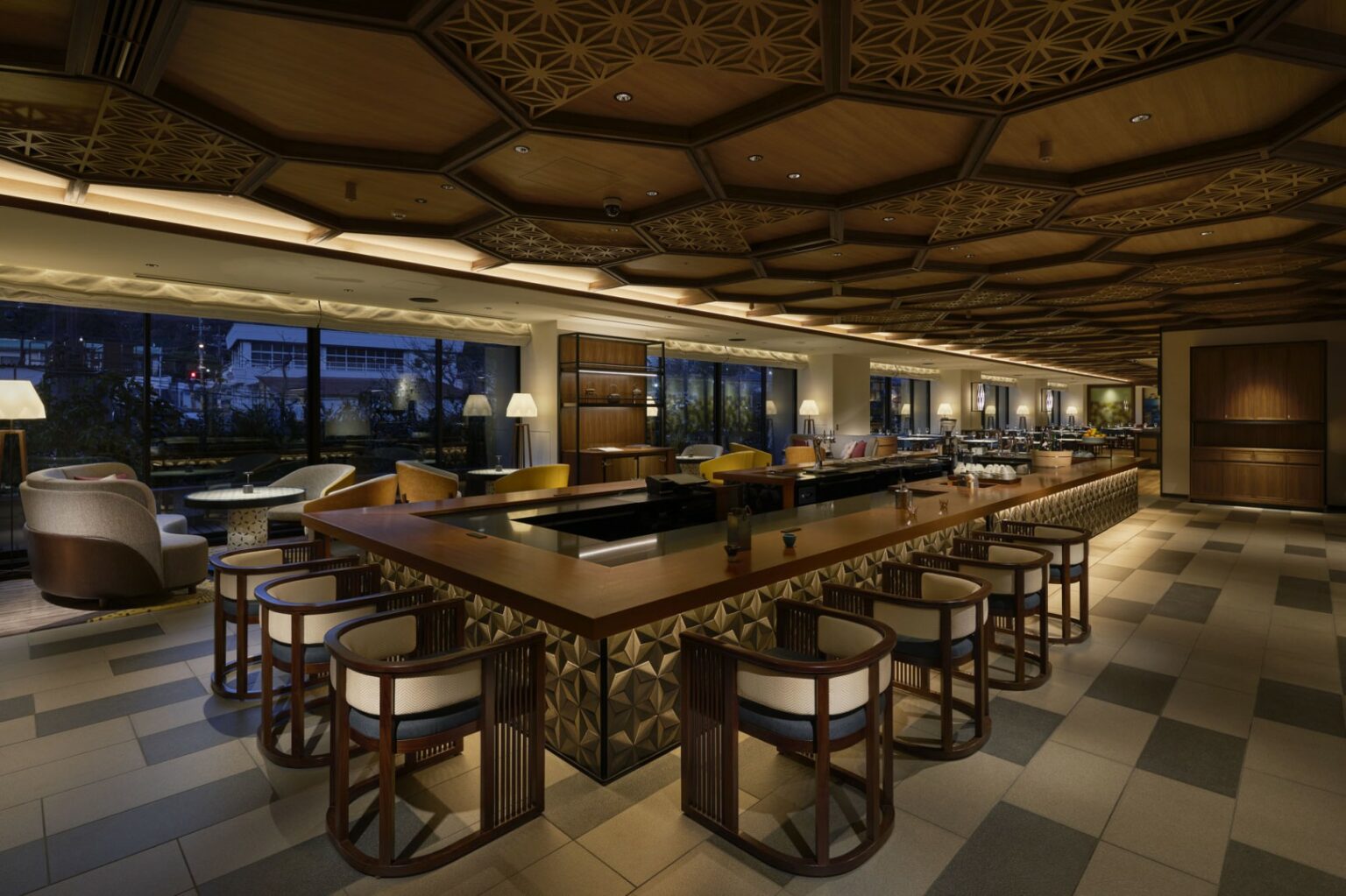 Modern restaurant lounge with warm lighting, featuring a large wooden bar, patterned ceiling, cushioned armchairs, and floor-to-ceiling windows at night. Cozy atmosphere.