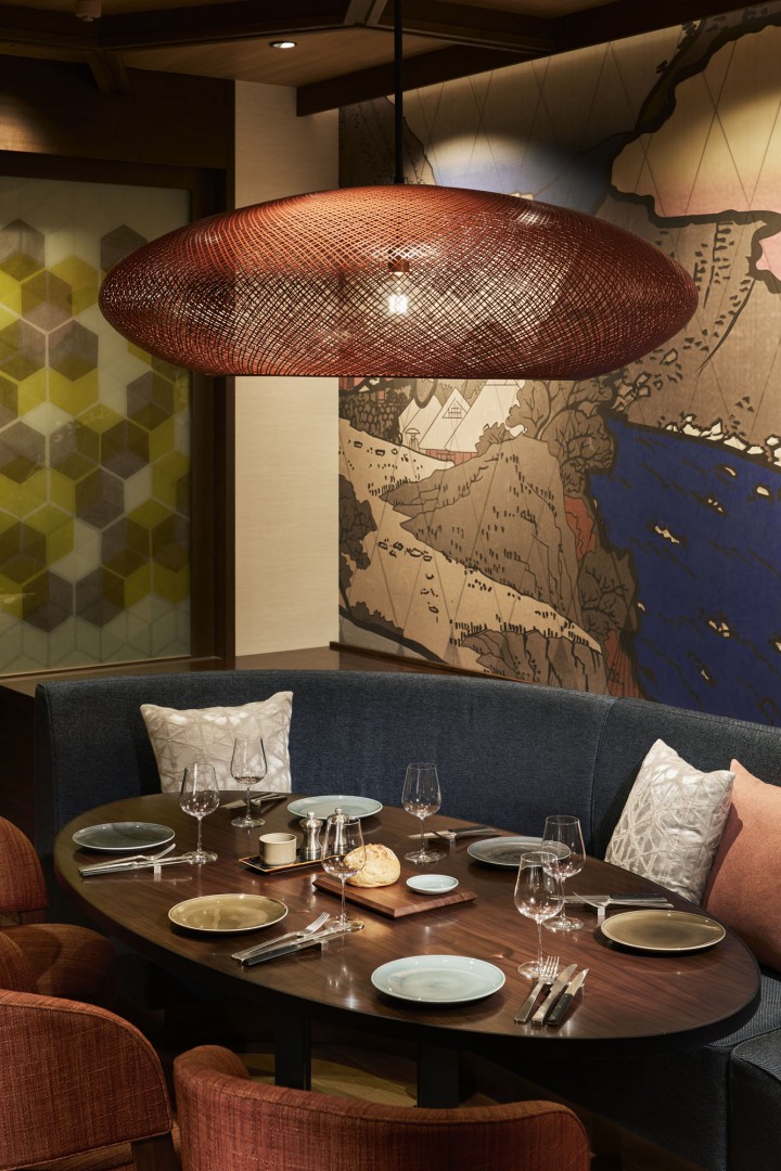 Modern dining area of a restaurant with a large oval wooden table set for six, featuring plates and glasses. Overhead, a stylish woven pendant light in warm tones. Behind, a mural with abstract landscape elements adds artistic flair. Cozy ambiance with cushioned chairs and pillows.
