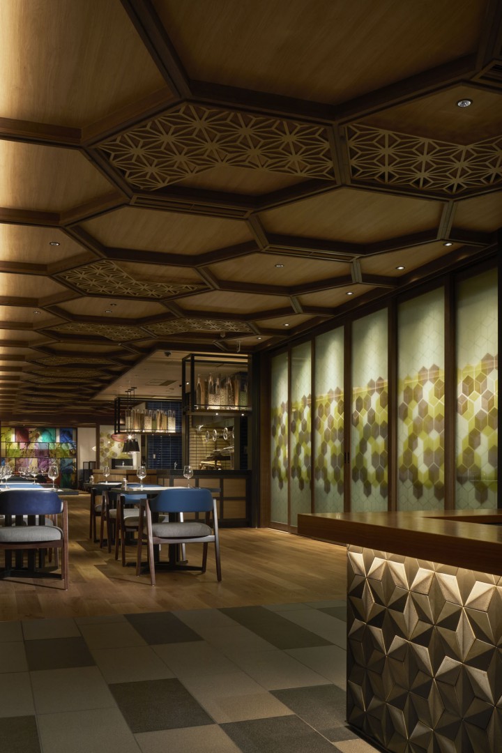 Modern restaurant interior with wooden hexagonal ceiling panels, geometric wall patterns, and ambient lighting. Stylish tables and chairs add elegance.