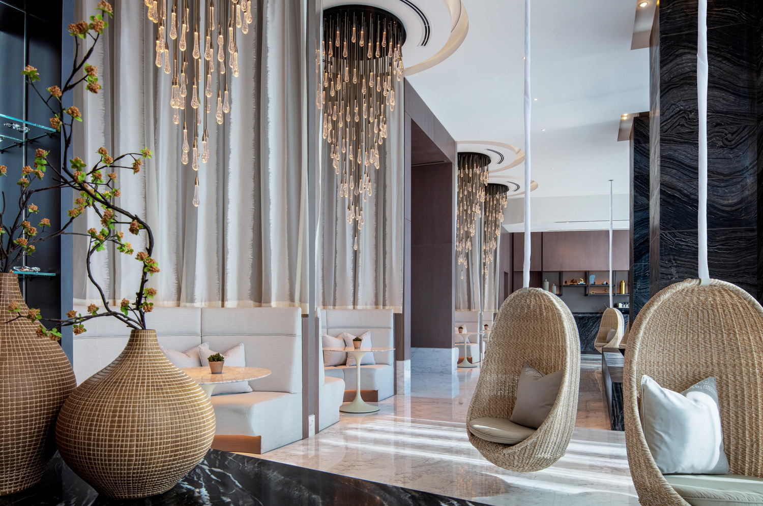 A chic spa reception area featuring woven chairs, elegant pendant lighting, and stylish decor, creating a warm and inviting ambiance for relaxation and socializing in a modern setting.