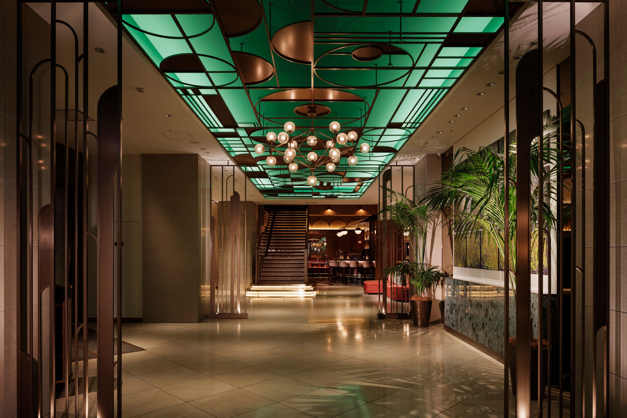 Luxurious hotel lobby with a green geometric ceiling, modern chandelier, and grand staircase. Surrounded by lush plants, creating a warm, inviting ambiance.