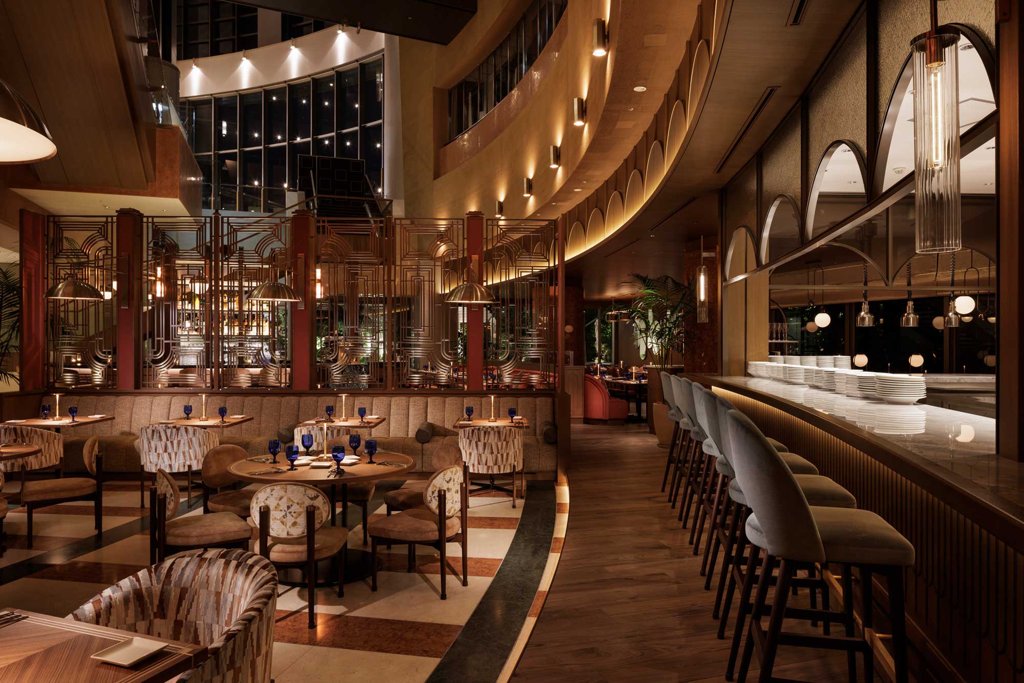 Elegant restaurant interior with warm lighting, featuring round wooden tables, upholstered chairs, and a sleek bar with high stools. Geometric designs create an art deco vibe.