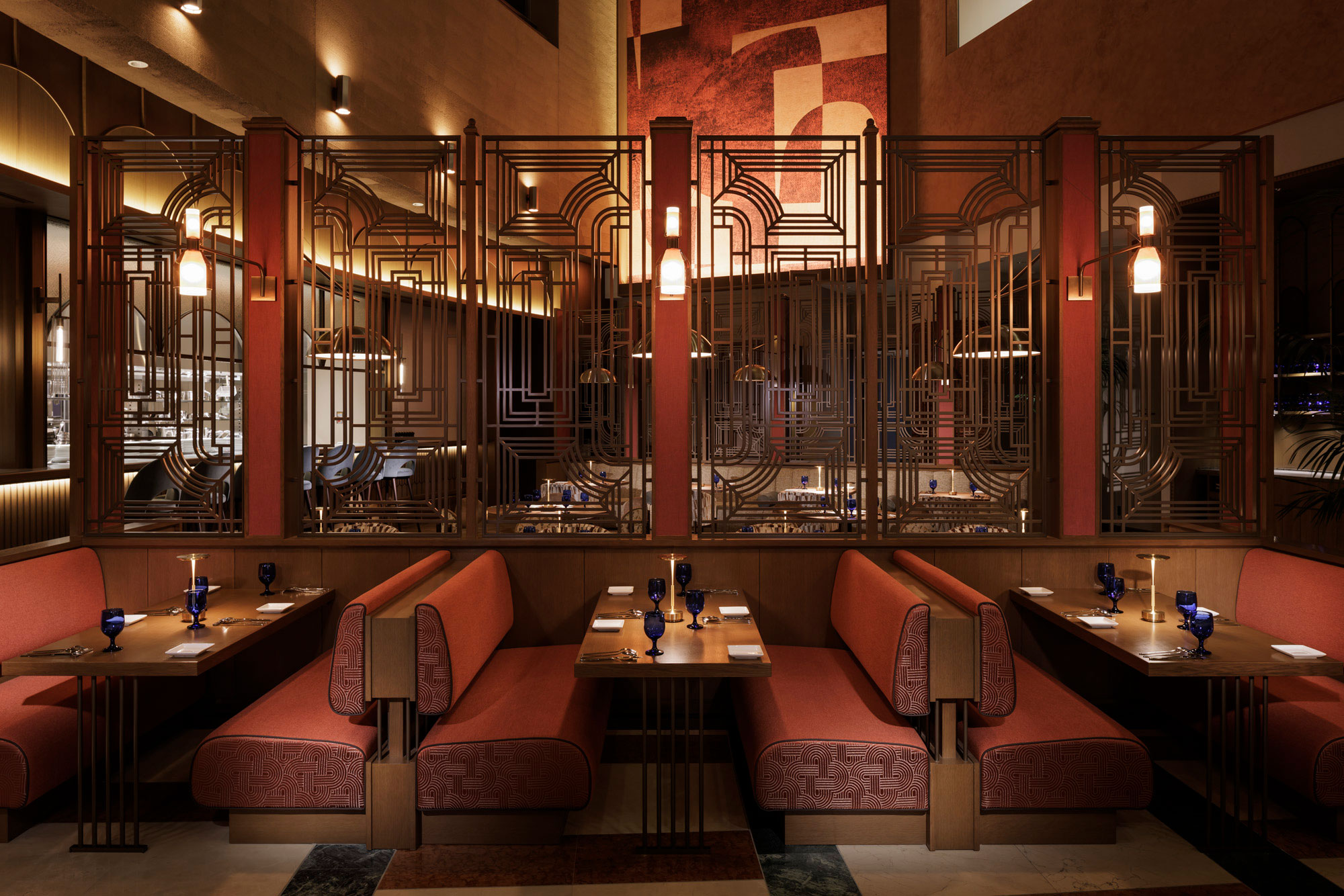 Art Deco-style restaurant interior with red cushioned booths, geometric metal dividers, warm lighting, and blue glassware. Elegant and inviting ambiance.