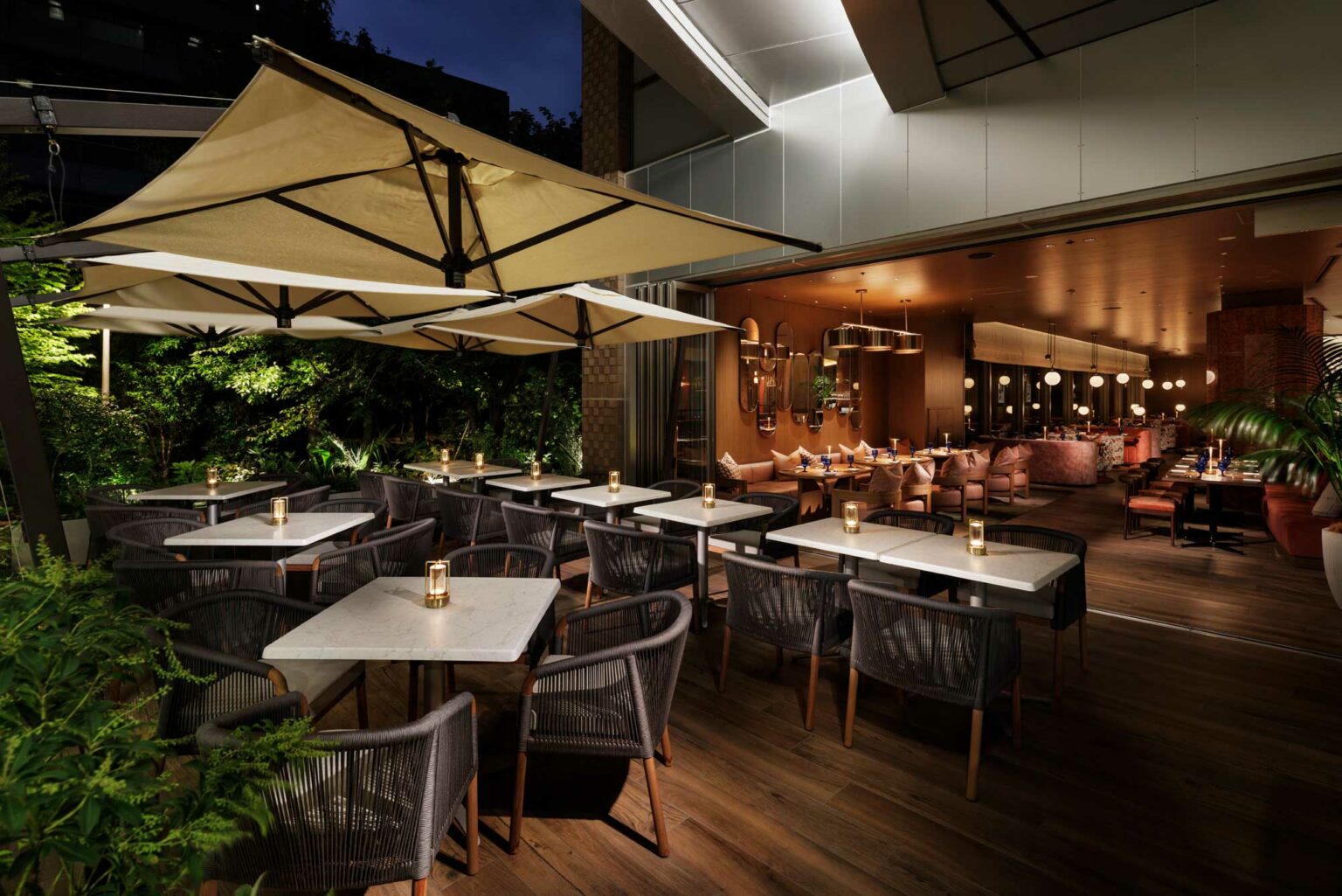 Chic outdoor and indoor dining area at night; white tables, wicker chairs, candlelit ambiance, large umbrellas, lush greenery, and warm interior lighting.