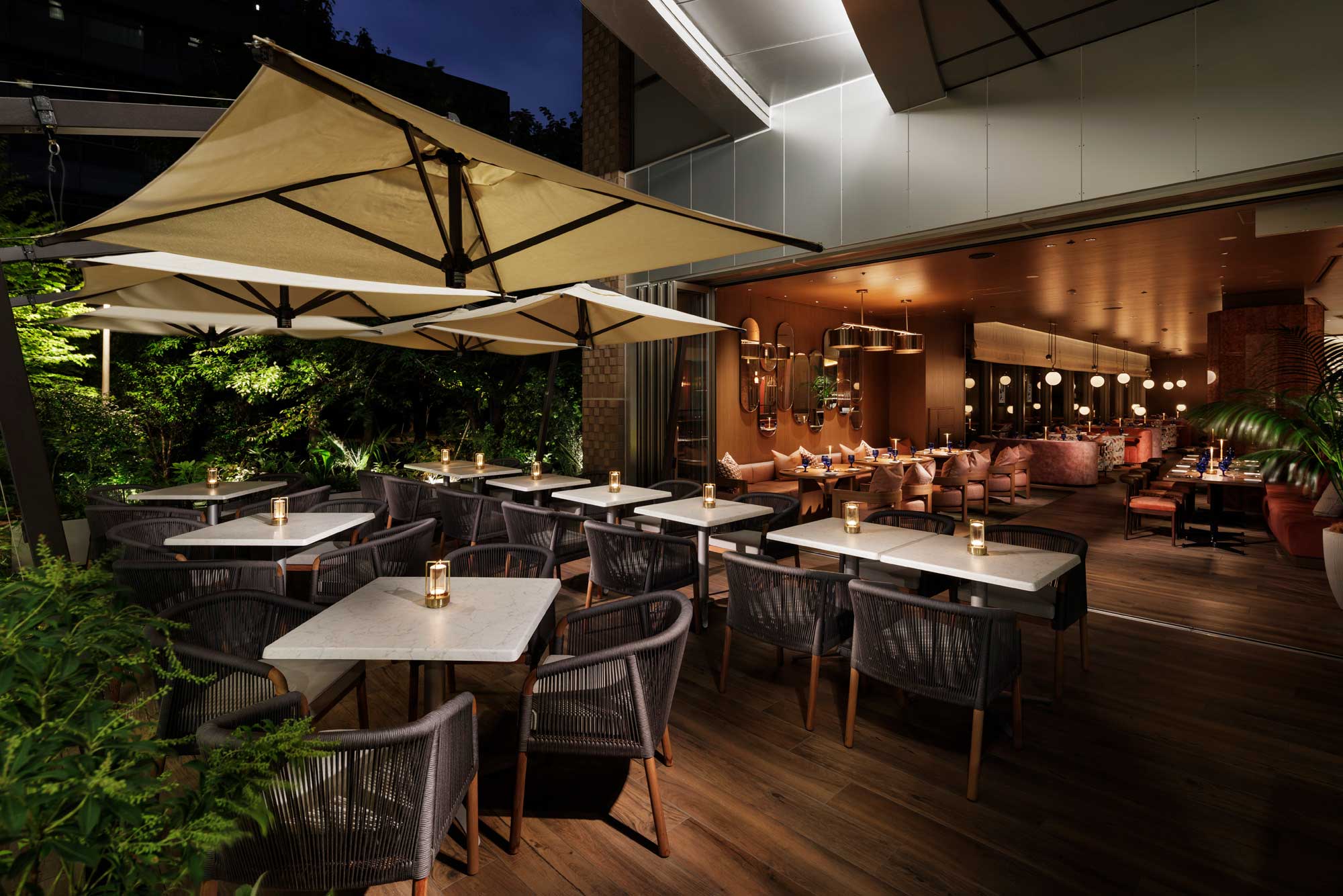 Chic outdoor and indoor dining area at night; white tables, wicker chairs, candlelit ambiance, large umbrellas, lush greenery, and warm interior lighting.