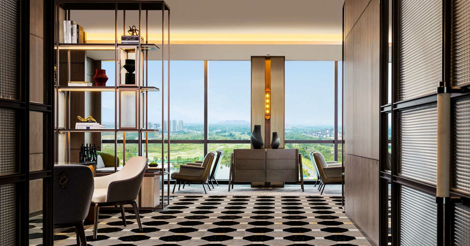 Sophisticated hotel lounge with modern furniture, large window offering a cityscape view, geometric patterned floor, and warm ambient lighting.
