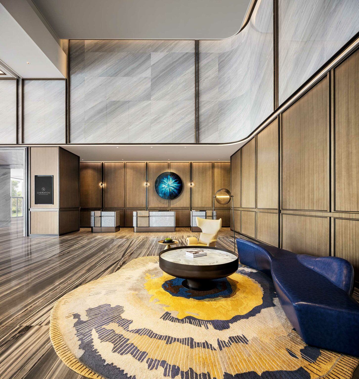 Spacious modern hotel lobby with high ceilings. Features a circular blue and yellow rug, dark blue curved sofa, round coffee table, and abstract wall art.