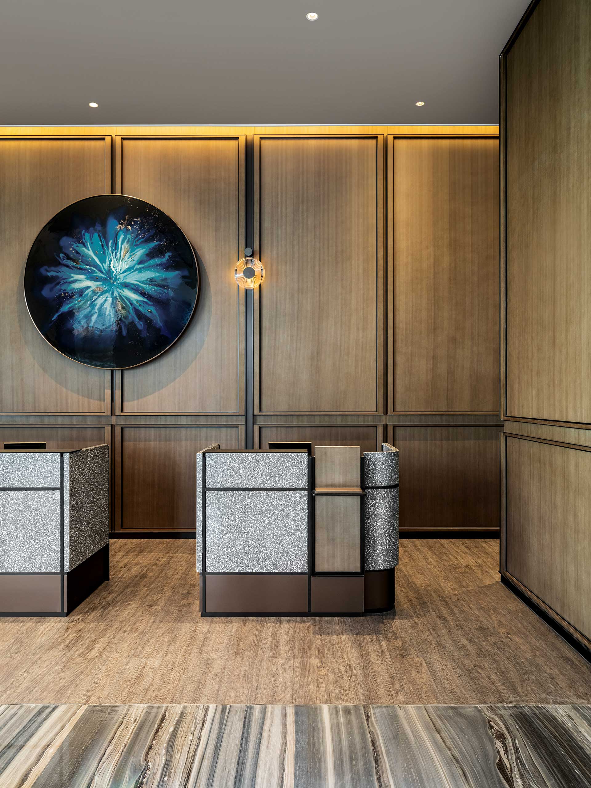 Modern hotel reception area with warm wood paneling, two sleek desks, and abstract blue artwork on the wall. Subtle lighting creates a calm atmosphere.