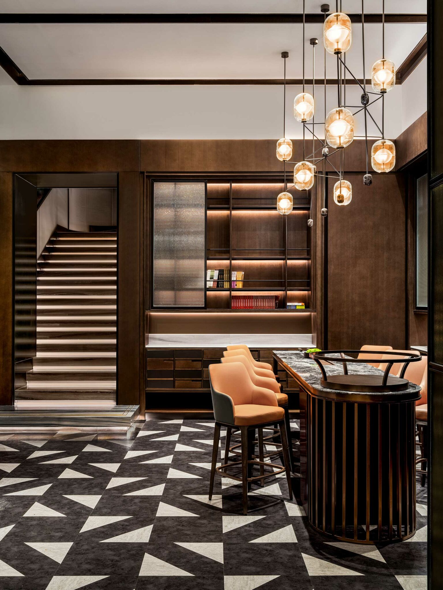 Modern hotel interior with a geometric patterned floor, warm lighting, and wood paneling. Features a bar with orange chairs and stairs leading upward.