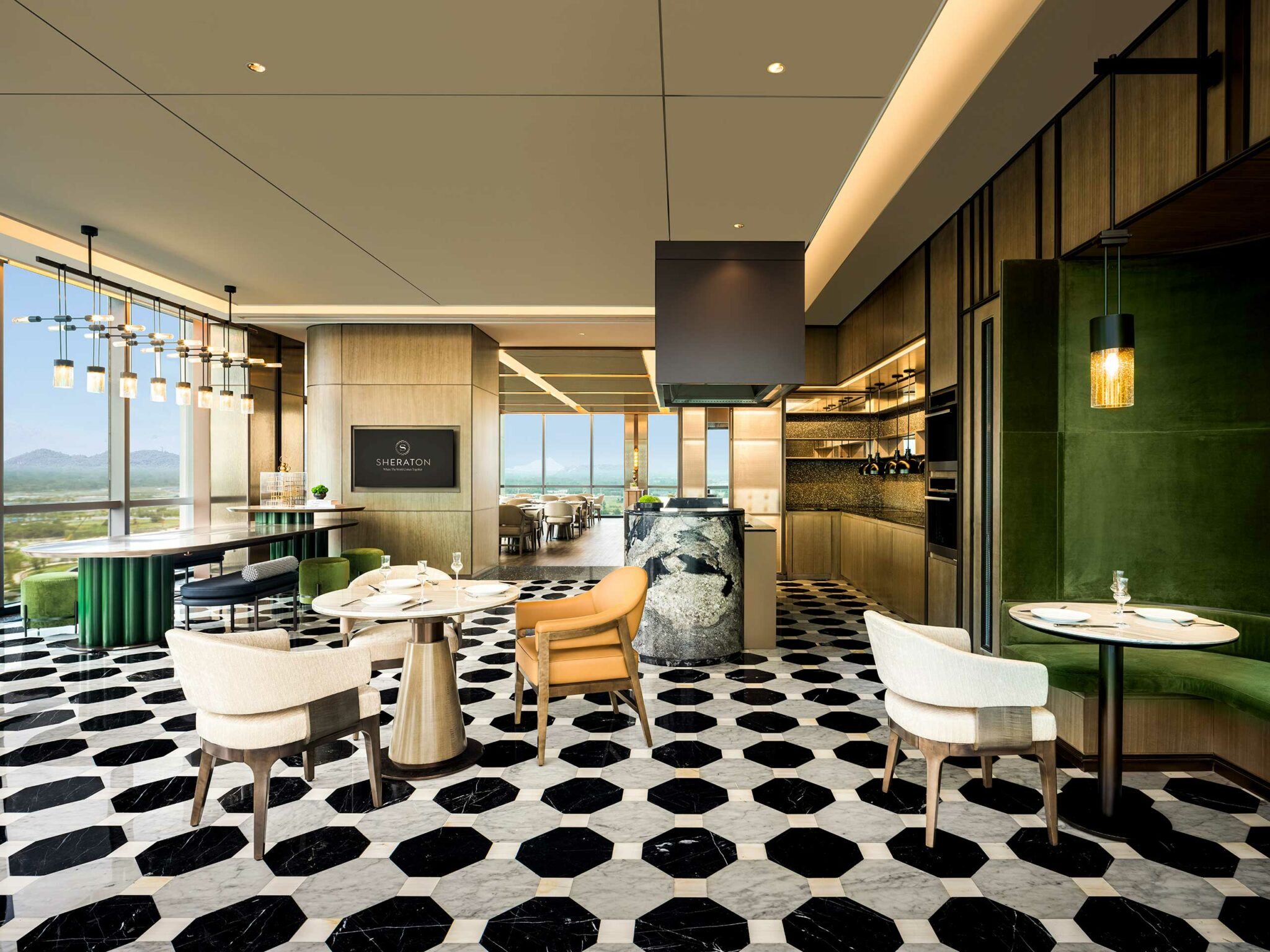 Modern hotel dining area with hexagonal black and white floor tiles, plush green and beige seating, soft pendant lighting, and large windows revealing mountain views.