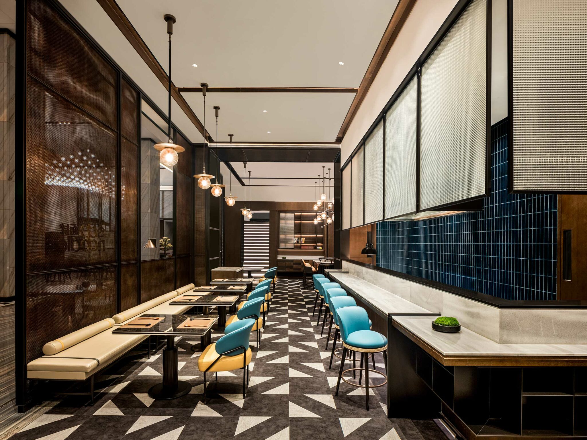 Modern, elegant hotel restaurant interior with geometric flooring, pendant lights, and aqua blue and yellow seating. The atmosphere is upscale and inviting.