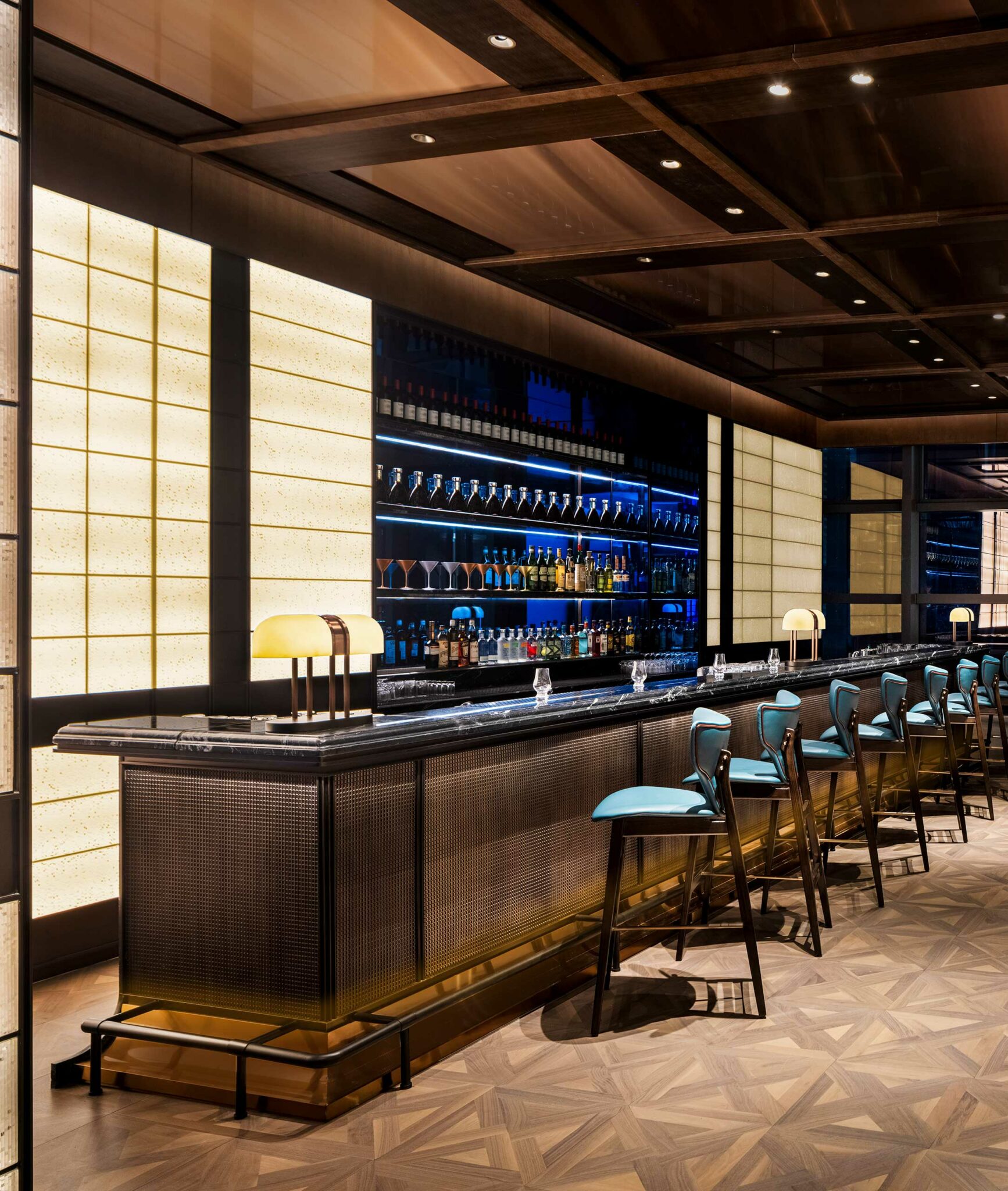 Sophisticated hotel bar with blue stools, elegant lighting, and wooden flooring. Seating includes modern chairs and round tables, set against a large window view.