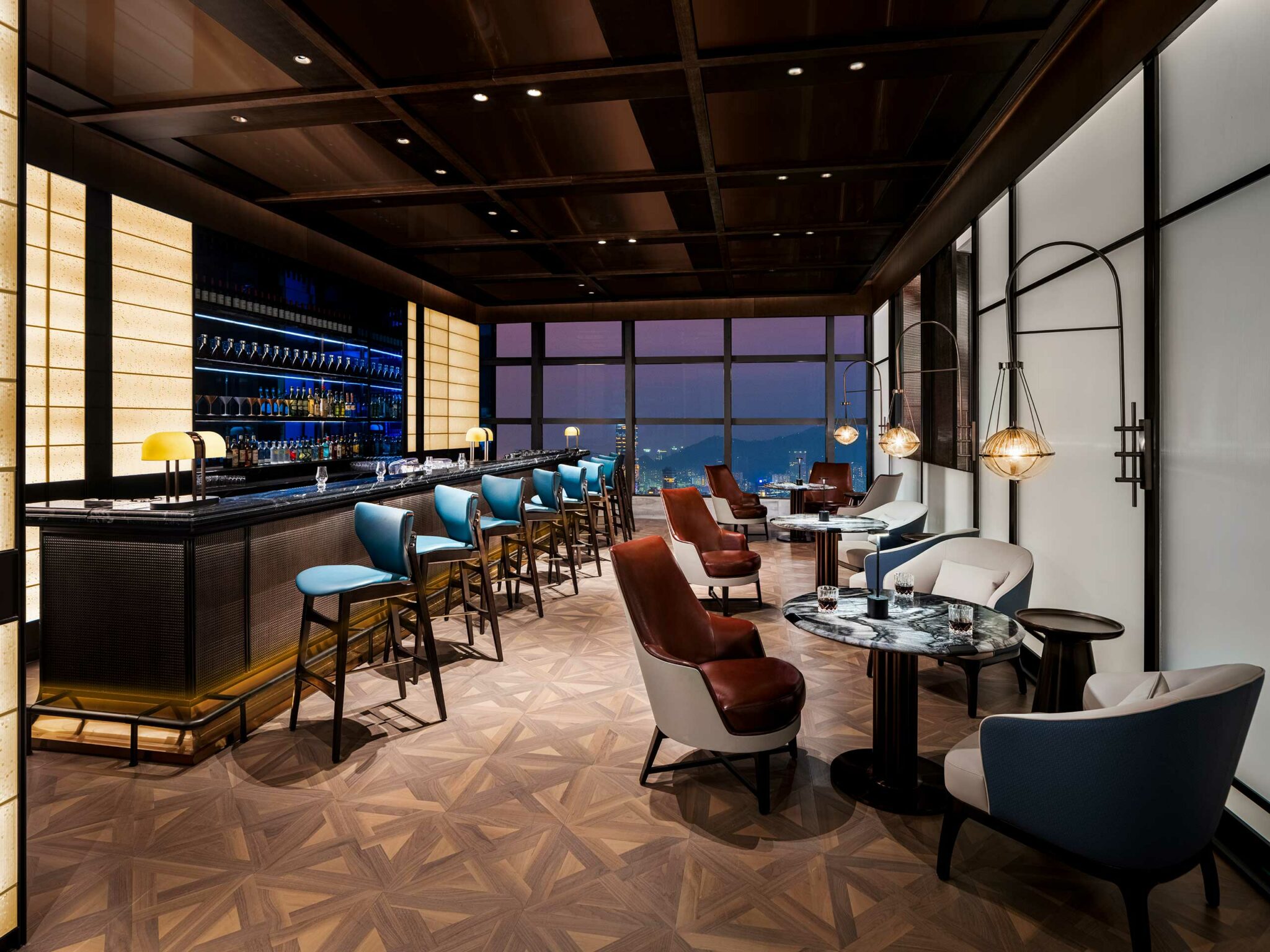 Sophisticated hotel bar with blue stools, elegant lighting, and wooden flooring. Seating includes modern chairs and round tables, set against a large window view.
