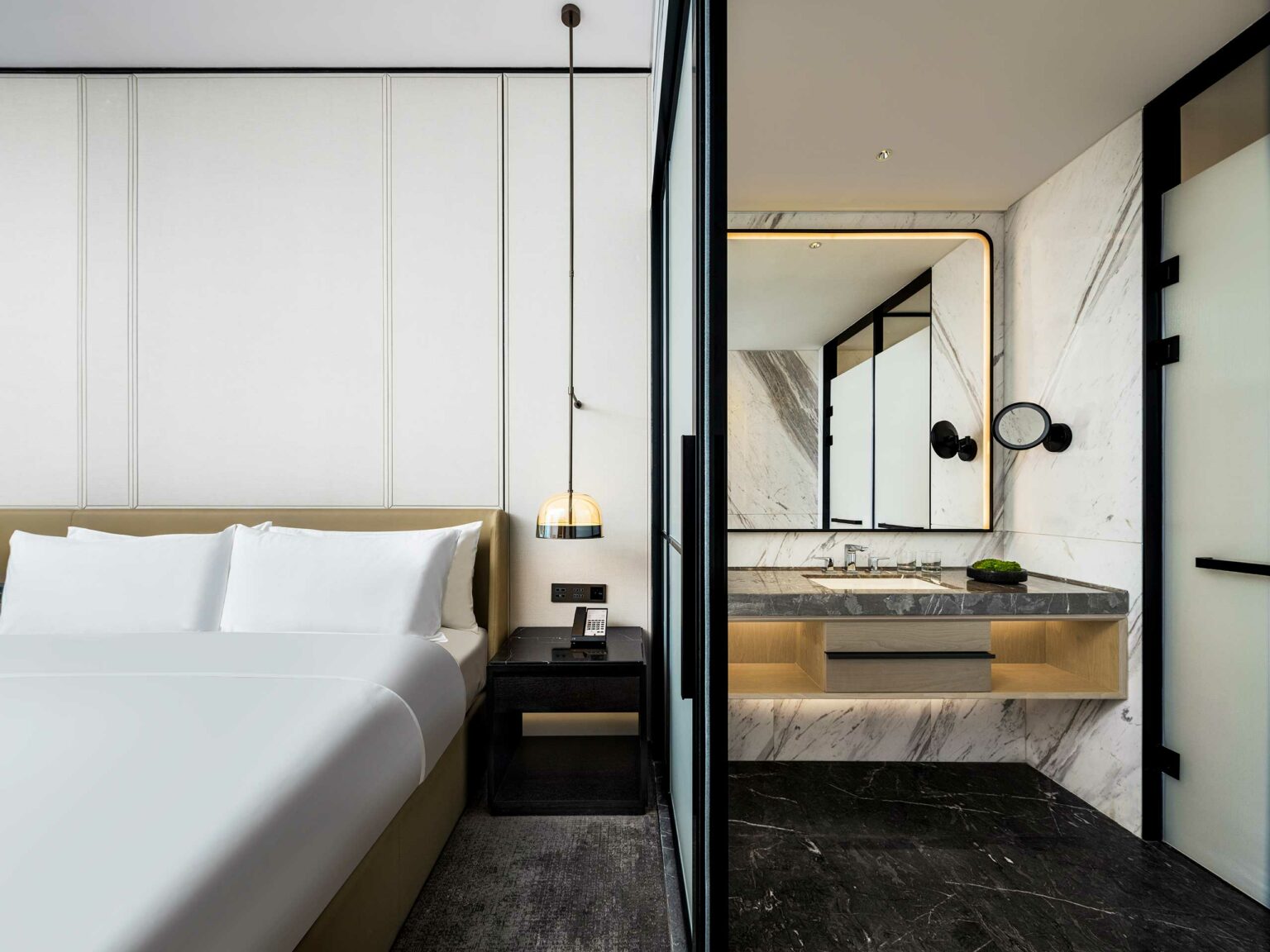 Modern hotel room featuring a white bed, sleek black nightstand with a phone, a stylish hanging lamp, and a marble bathroom with a large mirror.