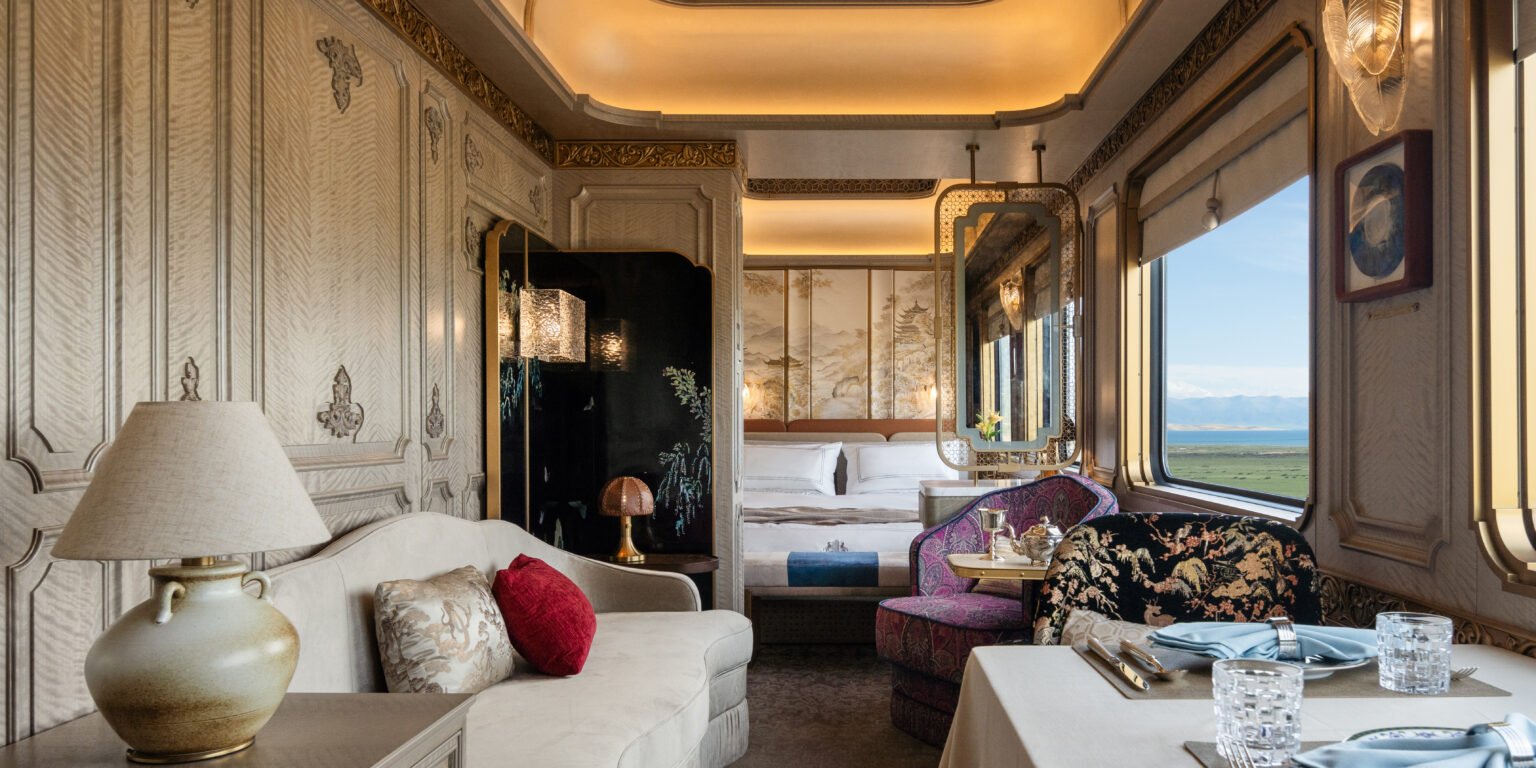 Luxurious train cabin with elegant decor, featuring ornate paneling, plush seating, a cozy bed, soft lighting, and scenic window view of rolling fields.