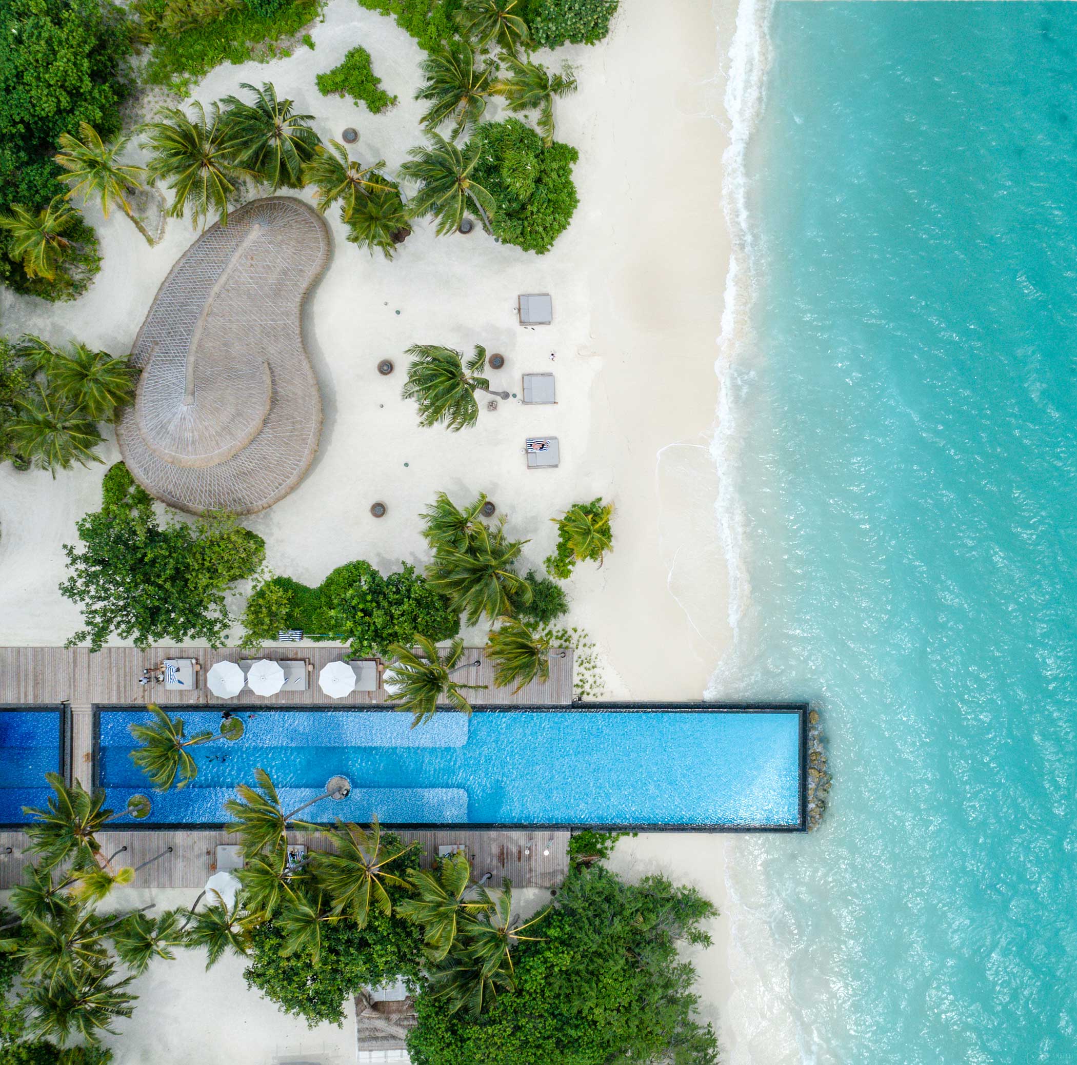 Aerial view of a tropical beach with turquoise water, lush greenery, and palm trees. A narrow pool and white umbrellas sit near the sandy shore.