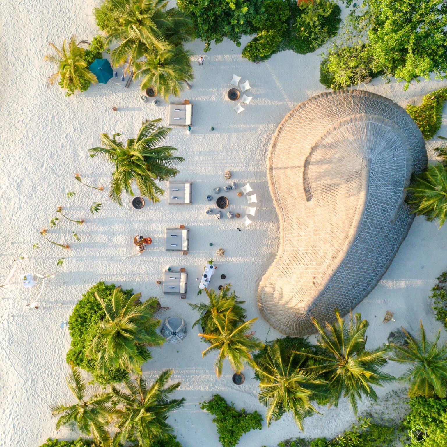 Aerial view of a serene beach setting with palm trees and a large, curved wicker structure on white sand. Lounge chairs and a table are arranged nearby.