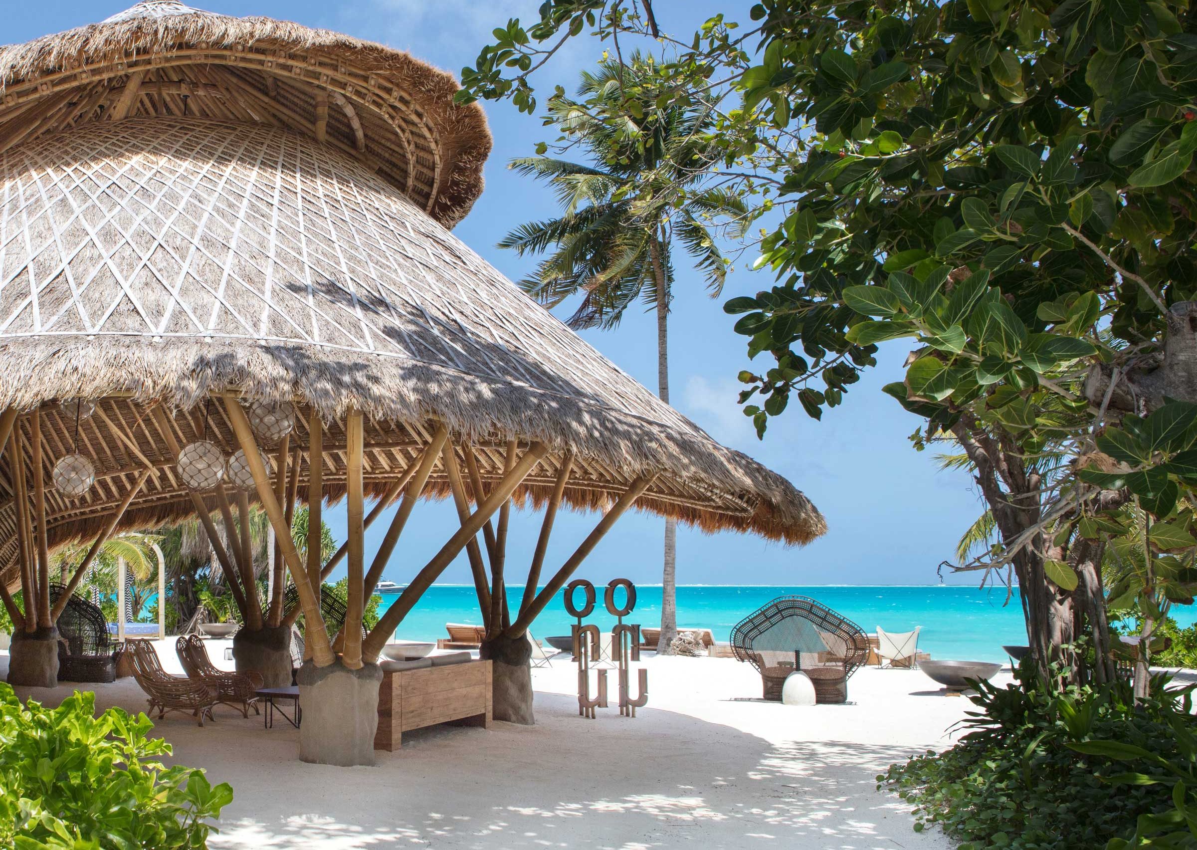 Tropical beach scene with a thatched-roof bamboo hut, palm trees, white sand, and turquoise ocean under a clear blue sky; serene and inviting atmosphere.