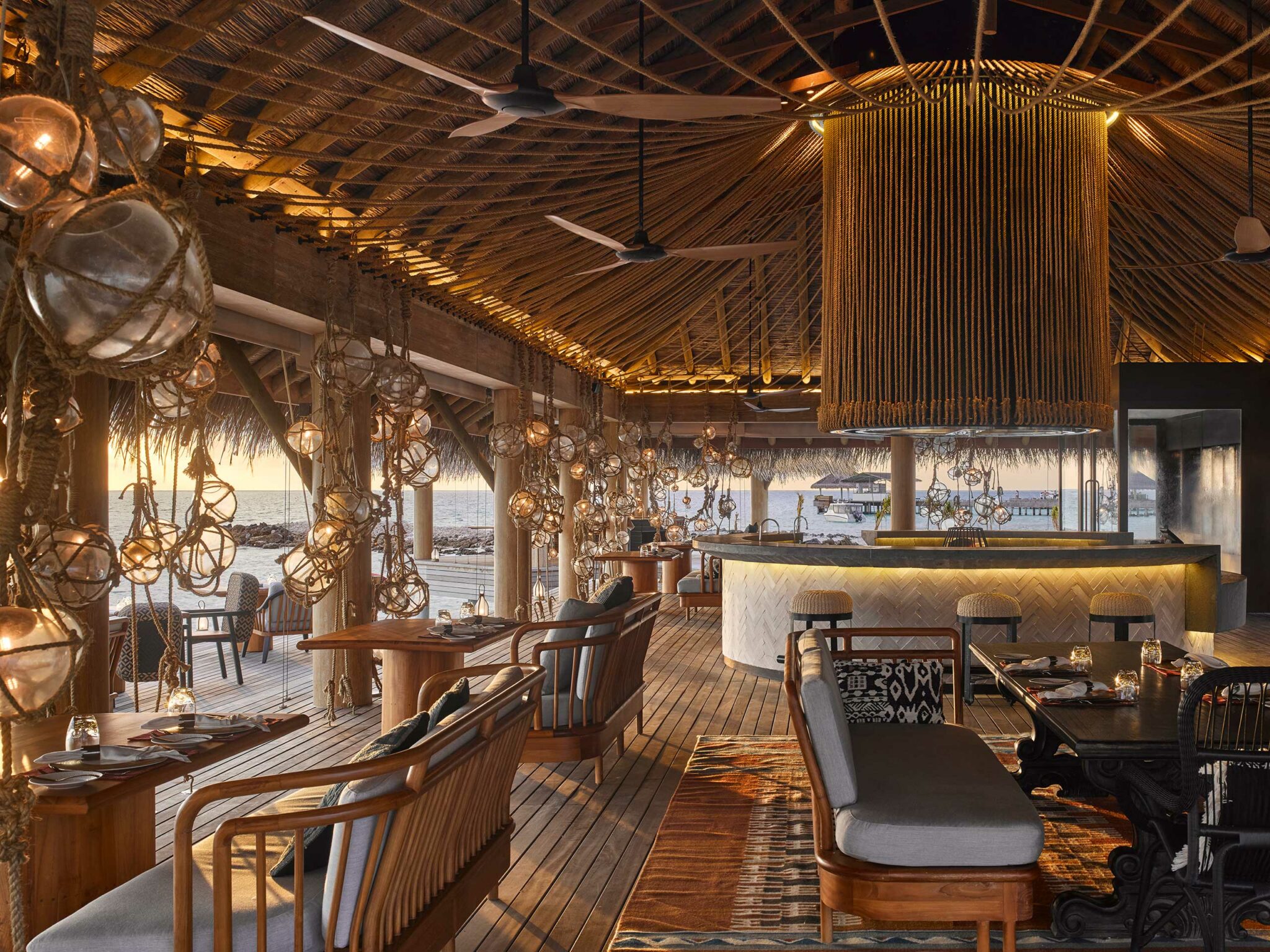 Cozy beachside resort restaurant interior with warm lighting, woven ceiling, and rustic decor. Open walls reveal an ocean view at sunset, creating a serene atmosphere.