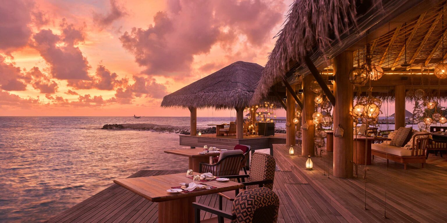 Tropical beachfront resort restaurant at sunset, with wooden tables and chairs, thatched roofs, and glowing lanterns reflecting on the calm ocean, creating a serene atmosphere.