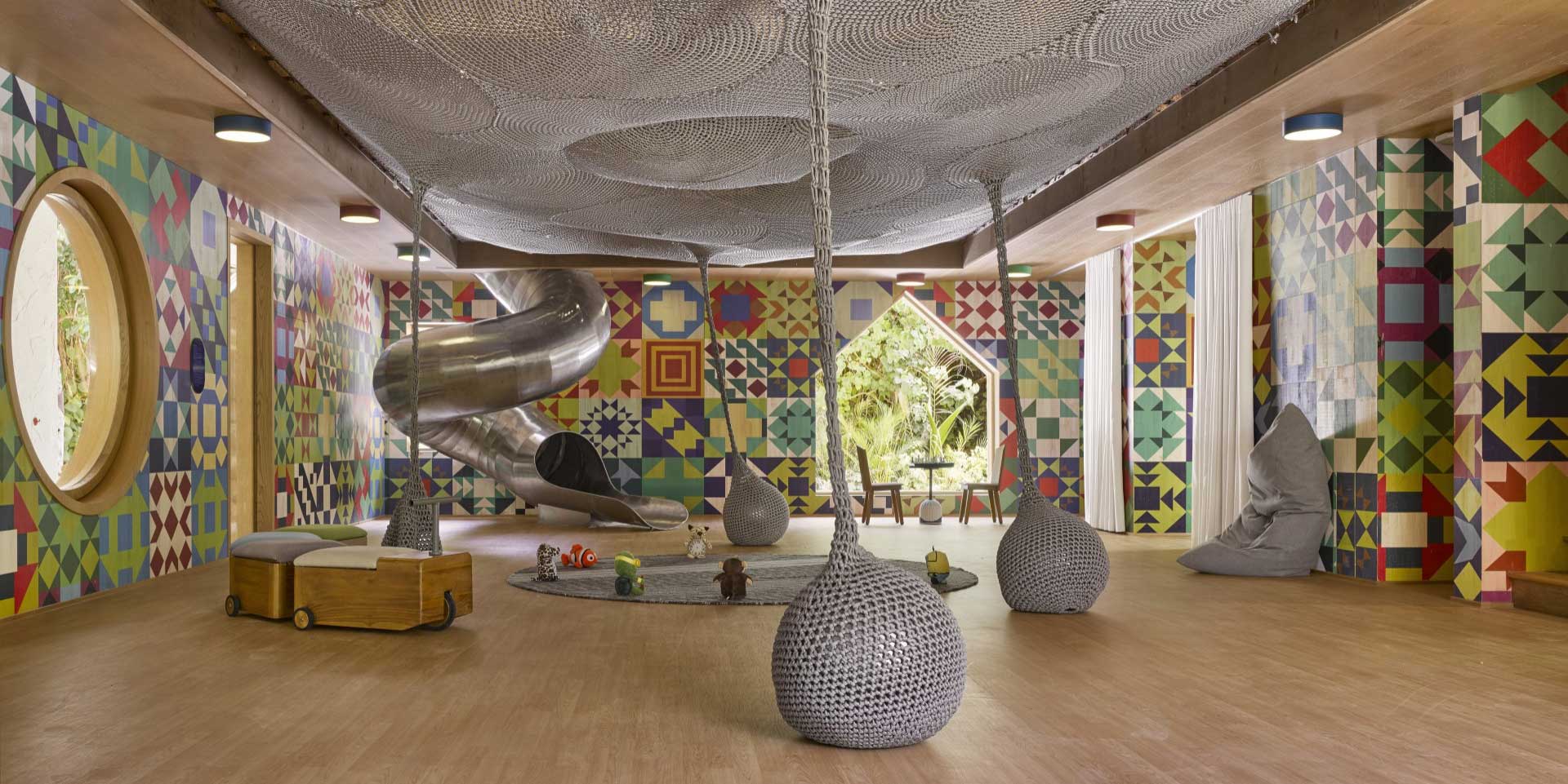 Vibrant resort playroom with colorful geometric walls, a spiral metal slide, ropes, and soft seating. Large windows frame lush greenery, creating a playful atmosphere.