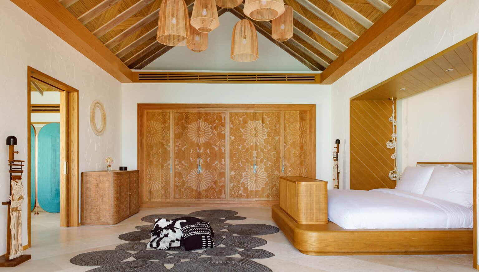 Spacious resort bedroom with wooden accents, a large bed, and wicker lamps. Floral-patterned wooden doors and a black-and-white rug add elegance. Relaxing ambiance.
