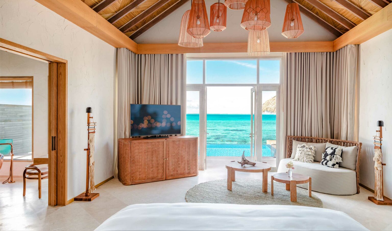 Luxurious beachfront resort room with ocean view. Features include a canopy bed, modern decor, flat-screen TV, wooden accents, and large windows. Relaxing and serene atmosphere.