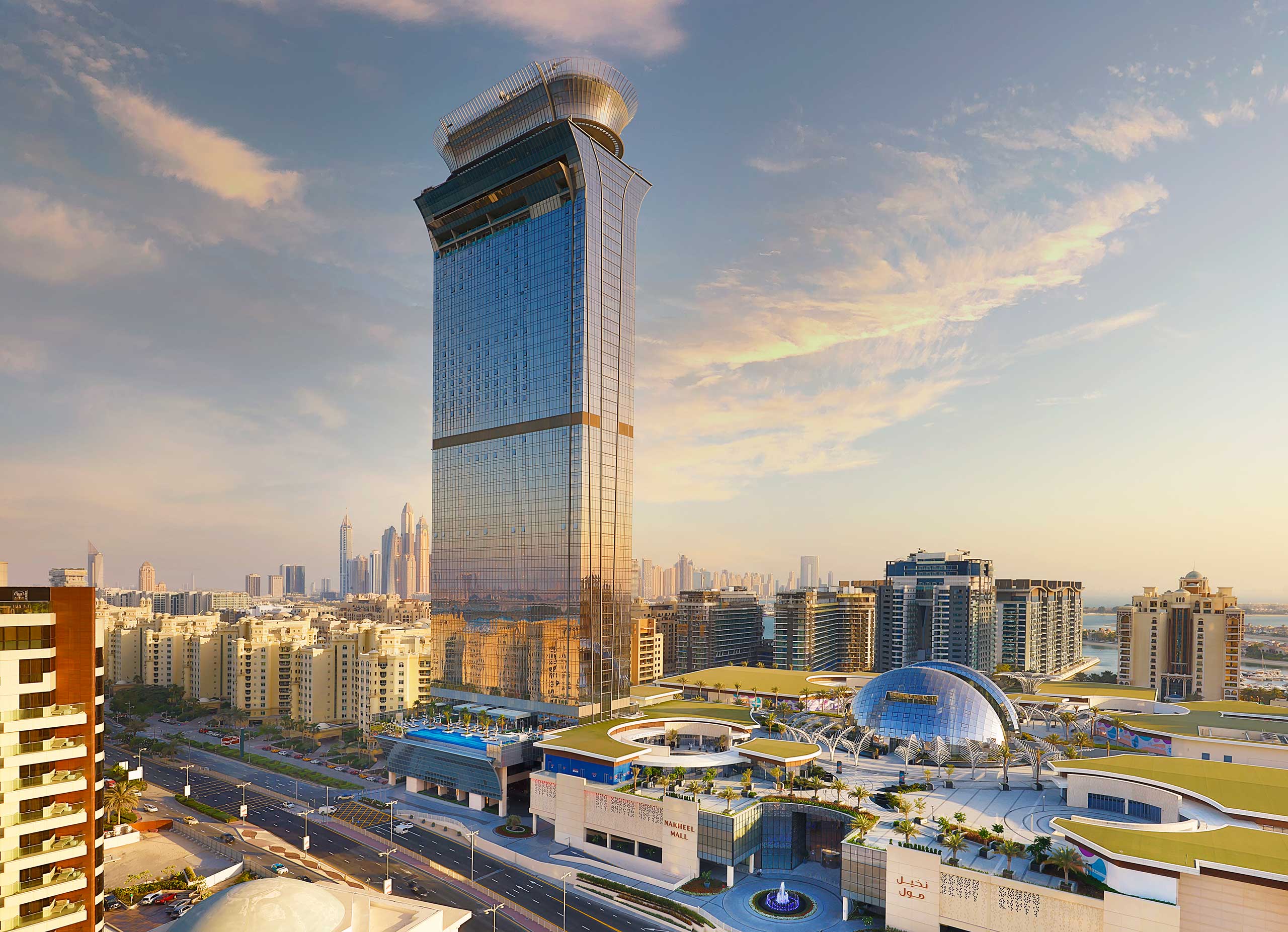 Exterior of The St. Regis The Palm, Dubai, featuring a tall, modern skyscraper with mirrored glass towers over Dubai's cityscape at sunset. Surrounding buildings and a domed structure are visible under a colorful sky.